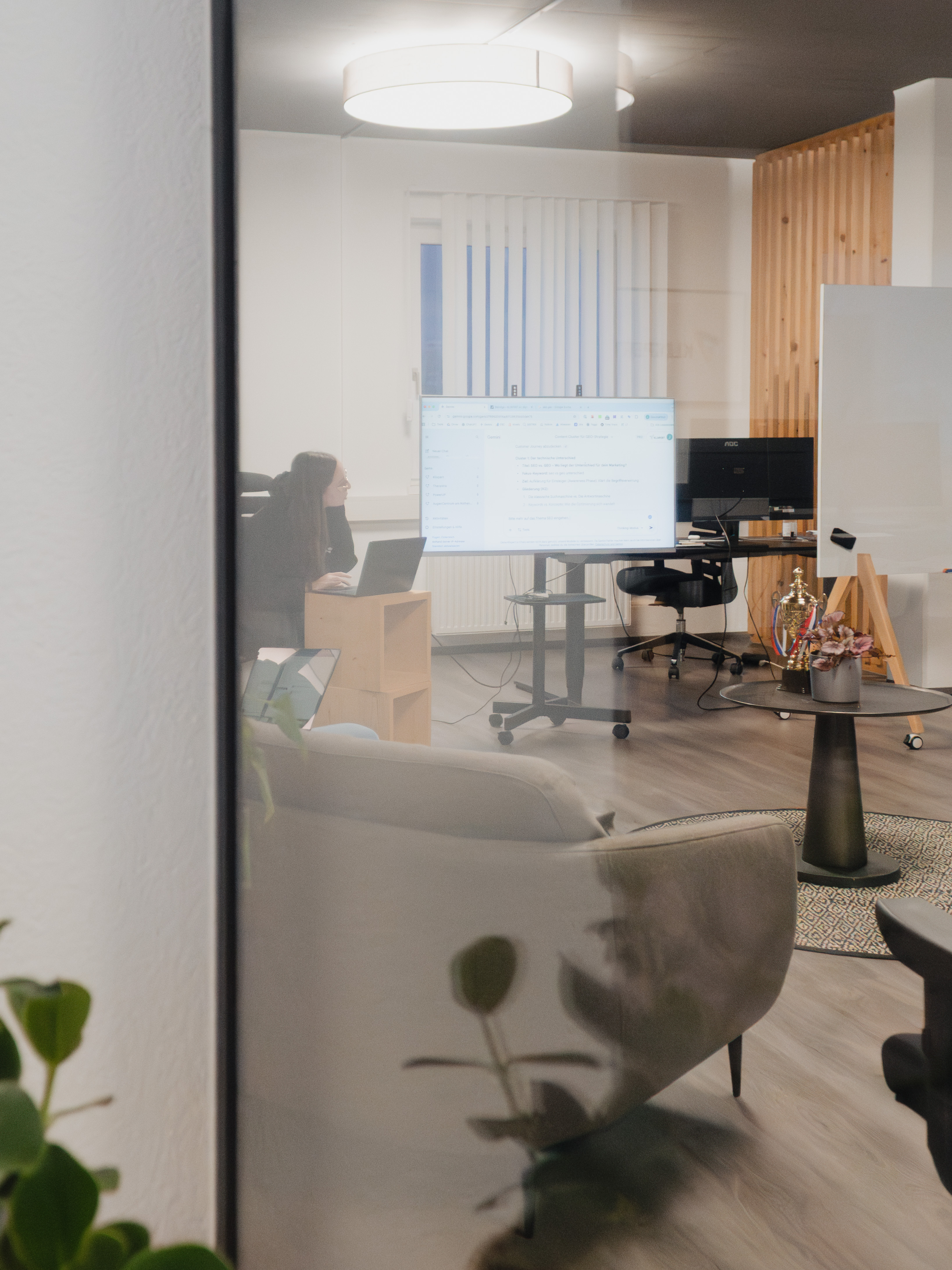 Blick durch eine Glaswand ins Büro: eine Person arbeitet am Laptop, daneben ein großer Bildschirm mit einem KI-Chatfenster.