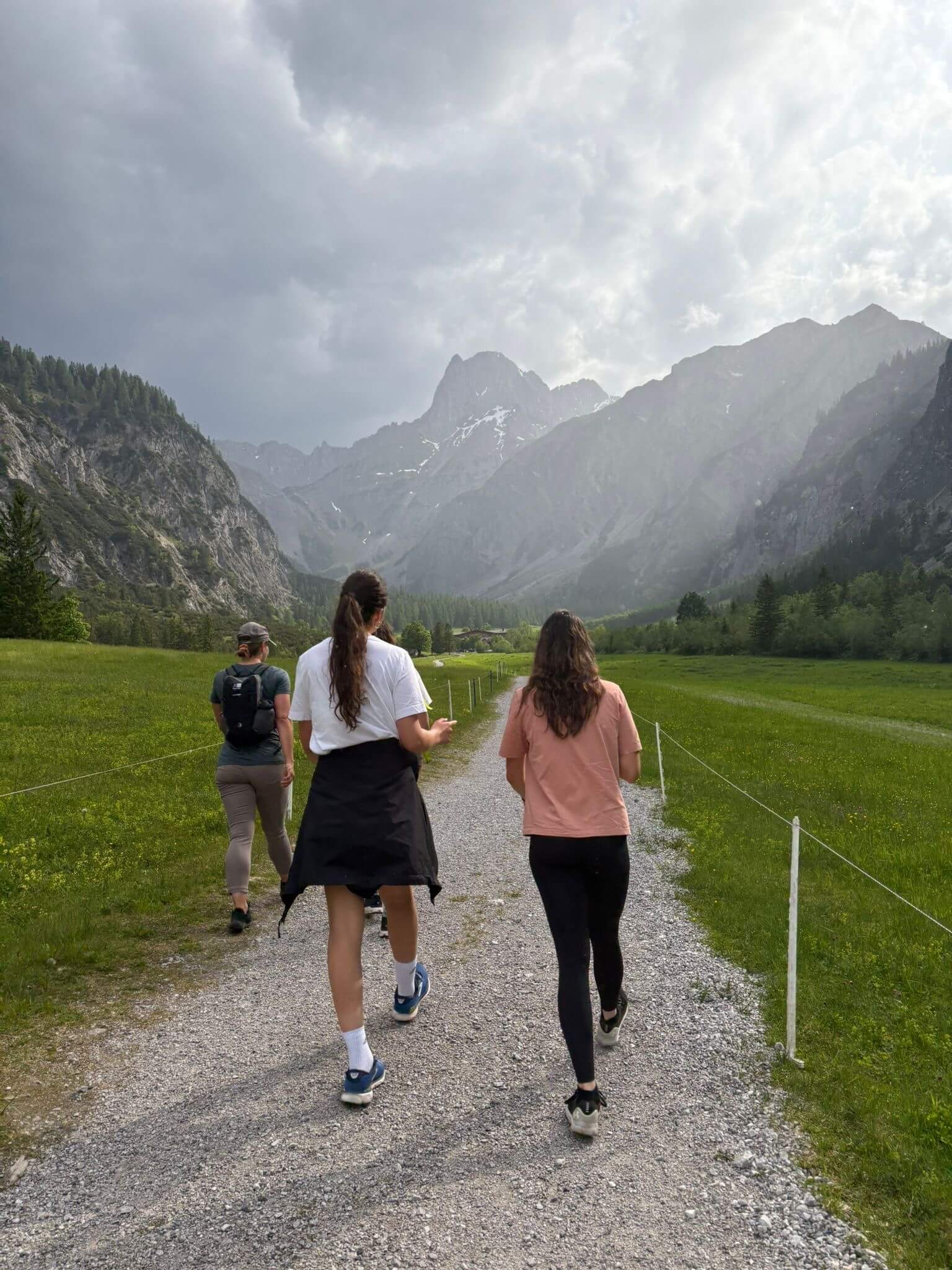 Drei Personen wandern auf einem Kiesweg durch eine grüne Berglandschaft, umgeben von hohen, wolkenverhangenen Gipfeln.