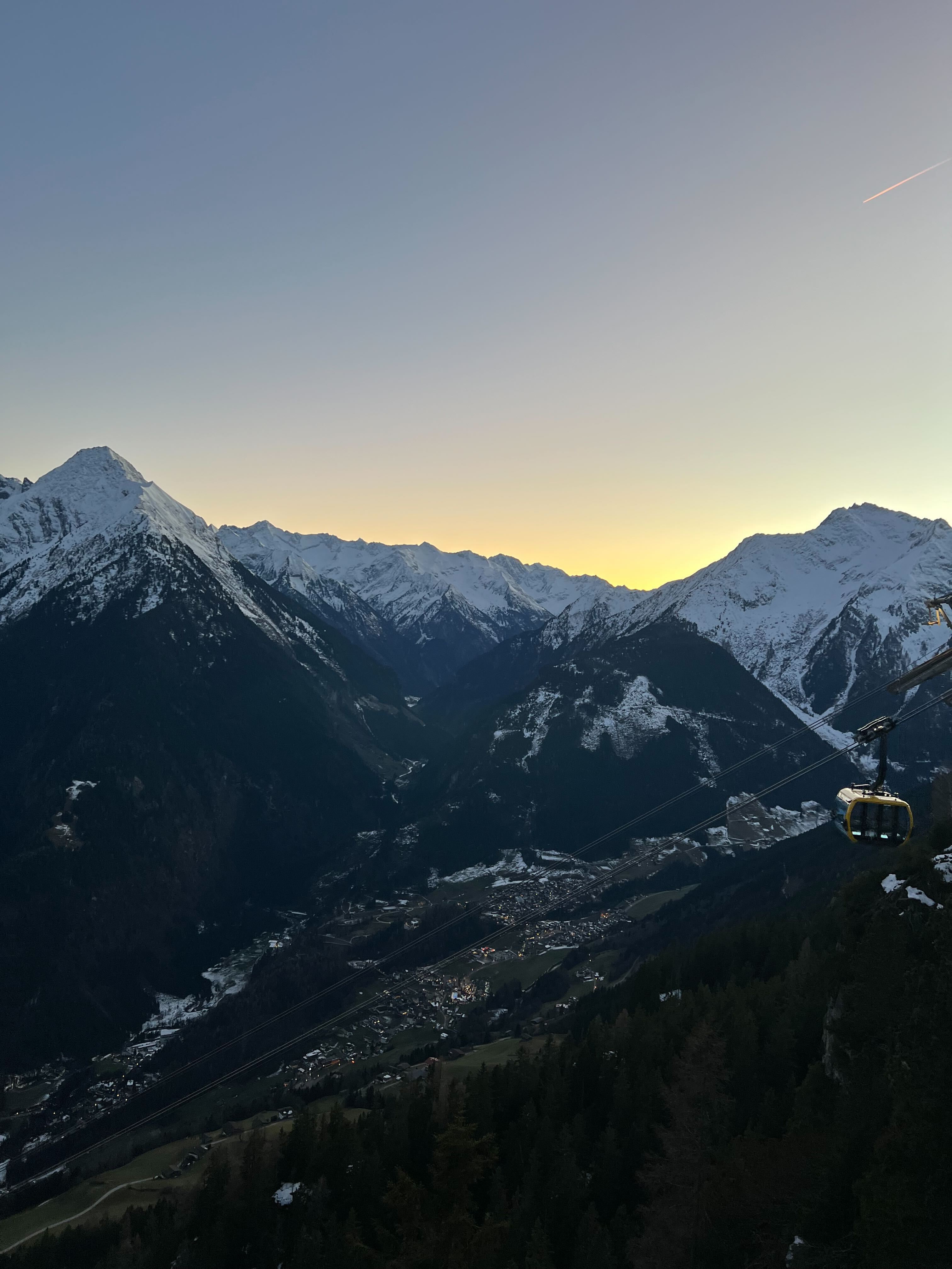 Blick von der Bergstation über das Tal: verschneite Gipfel im Abendlicht, eine Gondel schwebt über dem Hang.