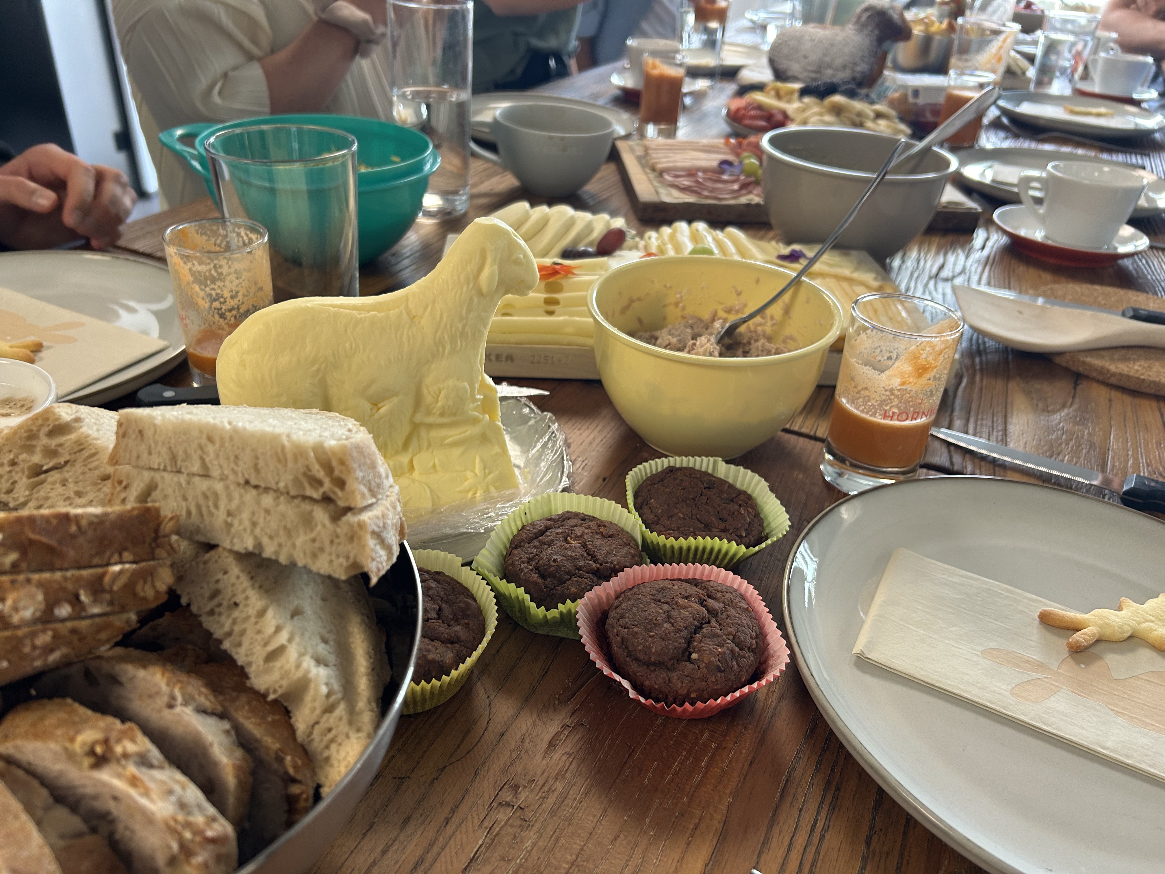 Frühstückstisch mit Brot, Schokoladenmuffins, Butter in Schaf-Form und verschiedenen Aufstrichen.