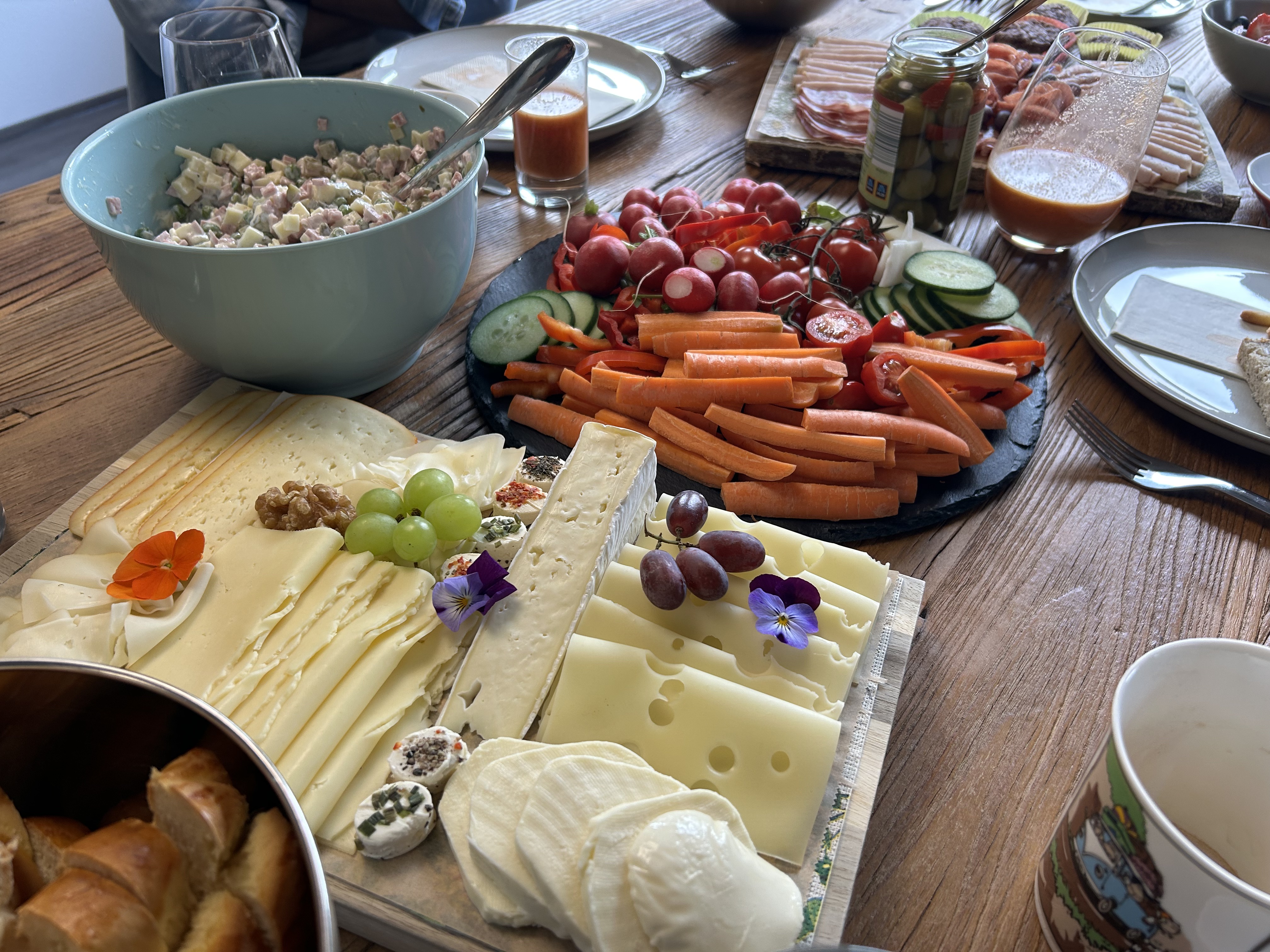 Vielfältiges Frühstücksbuffet mit Käseplatte, frischem Gemüse, Brotkorb und Salat auf Holztisch.
