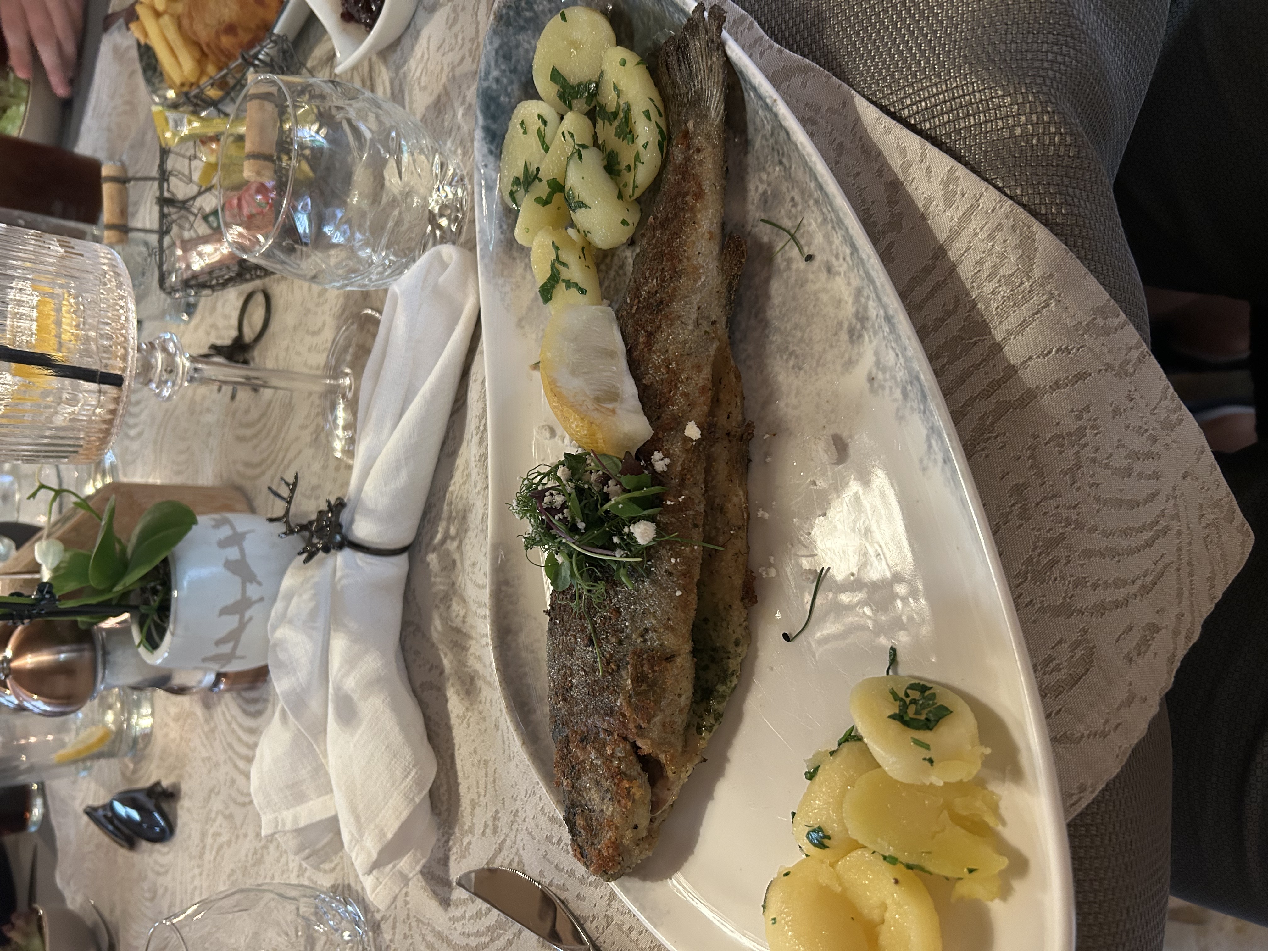Gebackener Fisch auf Teller mit Kartoffeln, garniert mit Kräutern, auf festlich gedecktem Tisch mit Besteck und Getränken.
