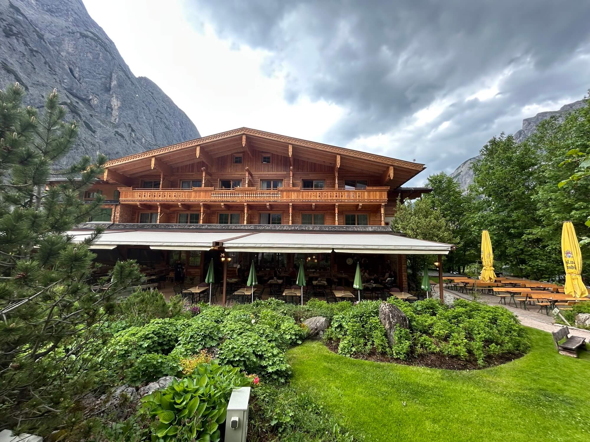 Alpenchalet mit Terrasse und Garten, umgeben von Bergen und Bäumen, unter bewölktem Himmel.
