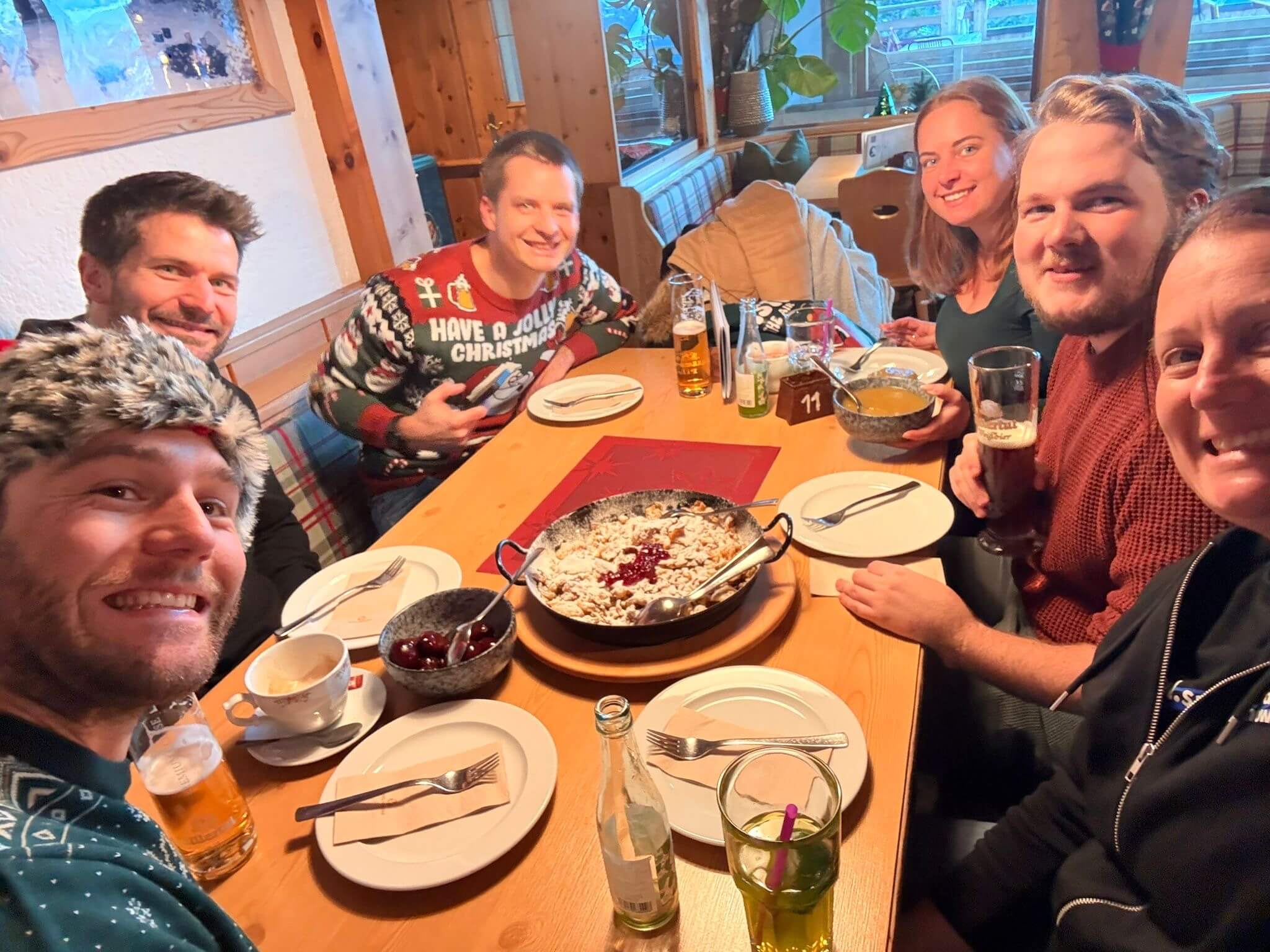 Sechs Personen um einen Tisch in der Almstube, in der Mitte eine Pfanne Kaiserschmarren mit Preiselbeeren.