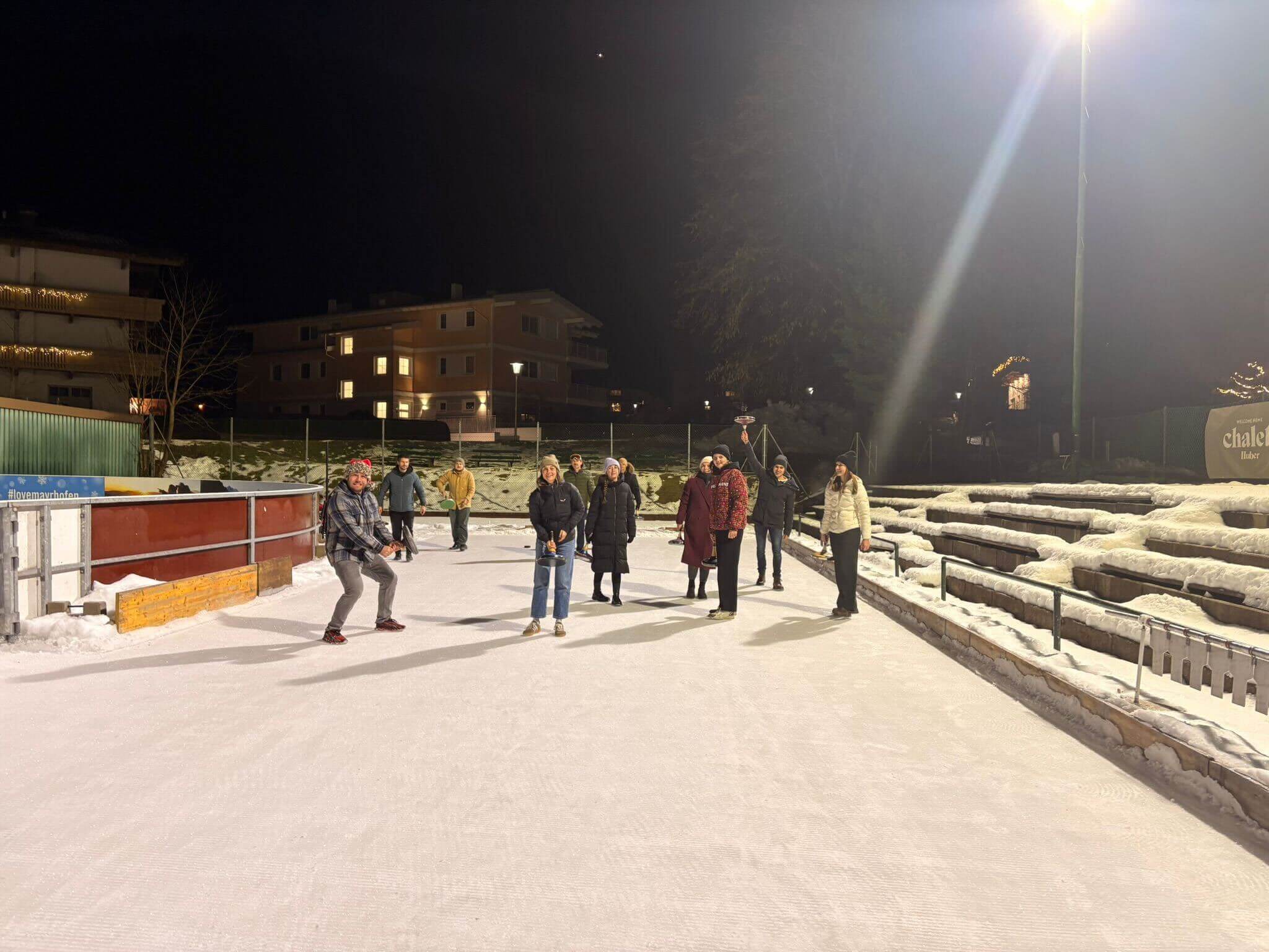 Die Gruppe verteilt sich unter Flutlicht auf der Eisbahn, dahinter verschneite Häuser und Zuschauerränge.