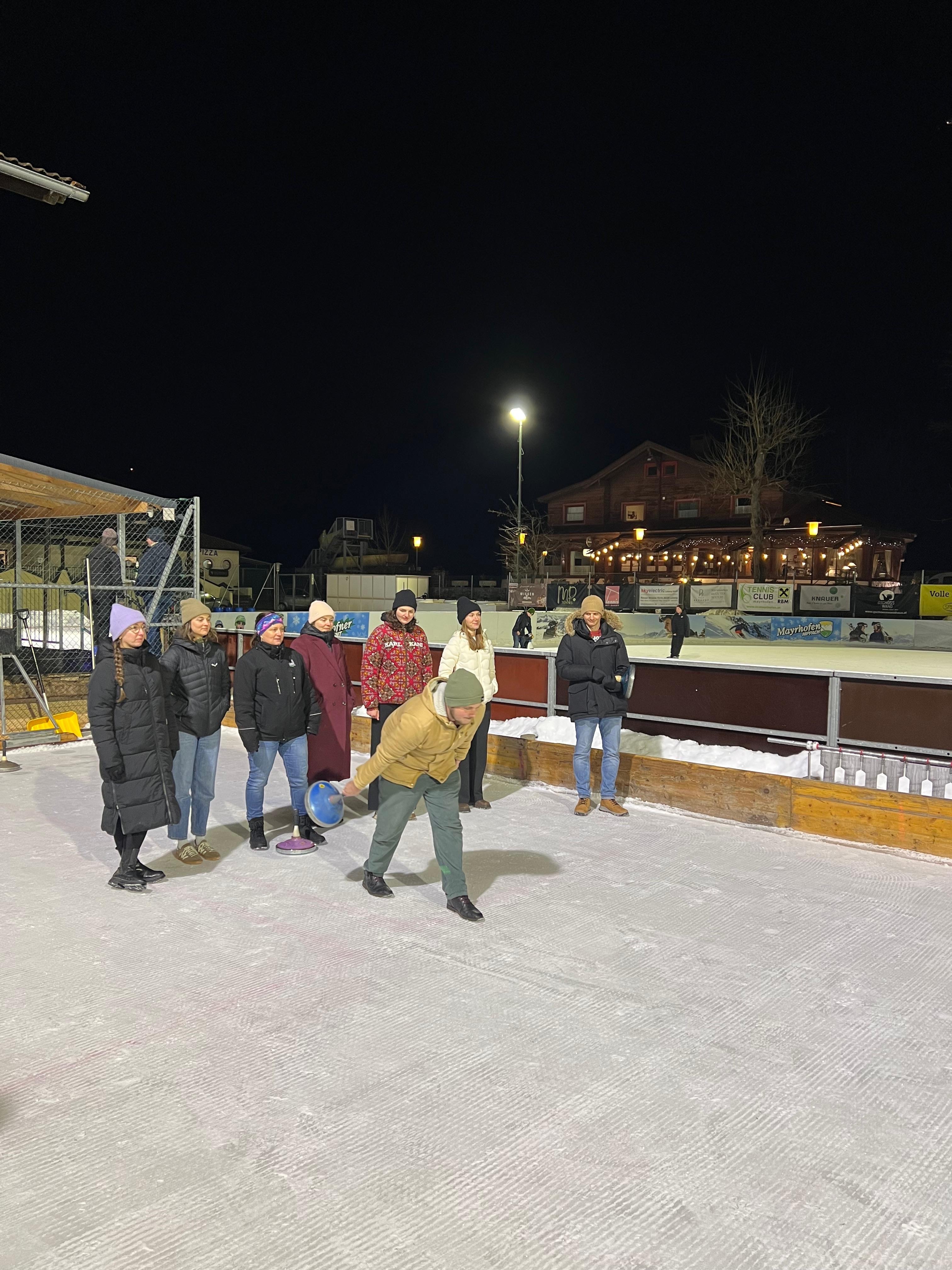 Person in gelber Jacke schiebt einen Eisstock an, am Rand der Eisbahn wartet das übrige Team.