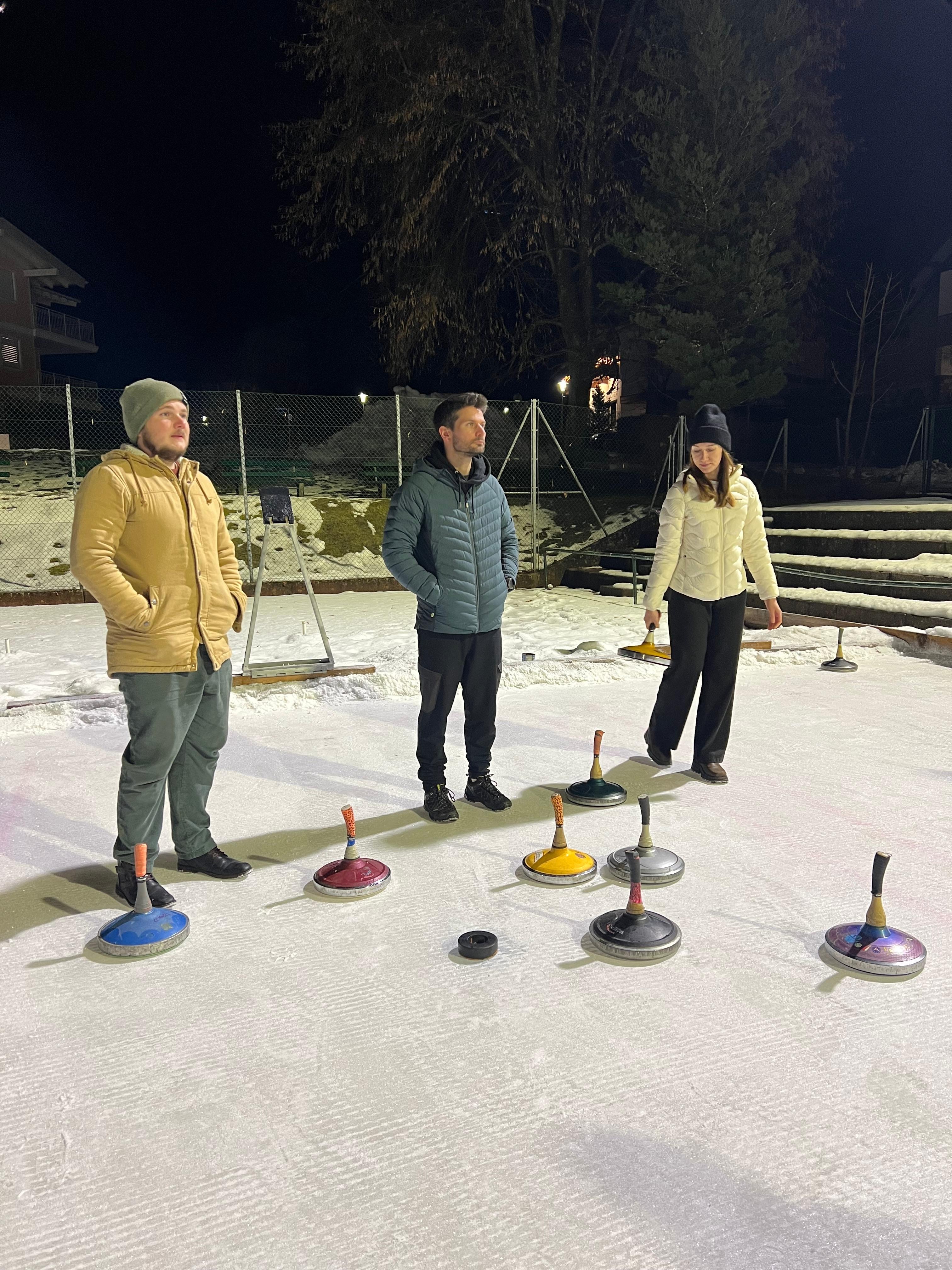 Drei Personen verfolgen den Lauf der bunten Eisstöcke, die über die Eisfläche verteilt liegen.
