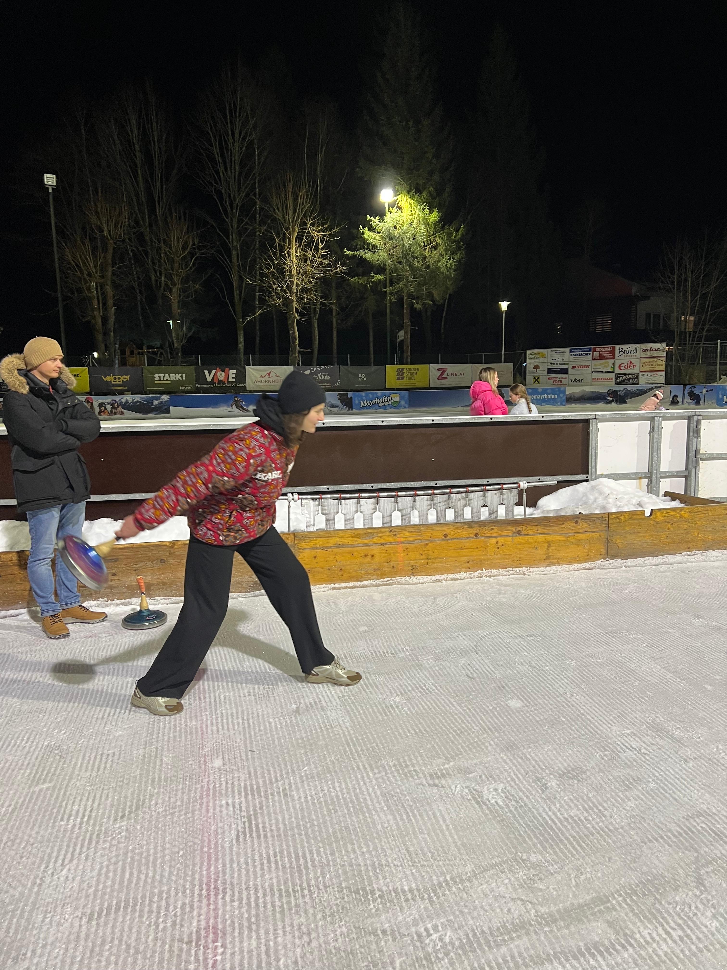 Frau in rot gemusterter Jacke holt zum Wurf mit dem Eisstock aus, auf der flutlichtbeleuchteten Eisbahn bei Nacht.