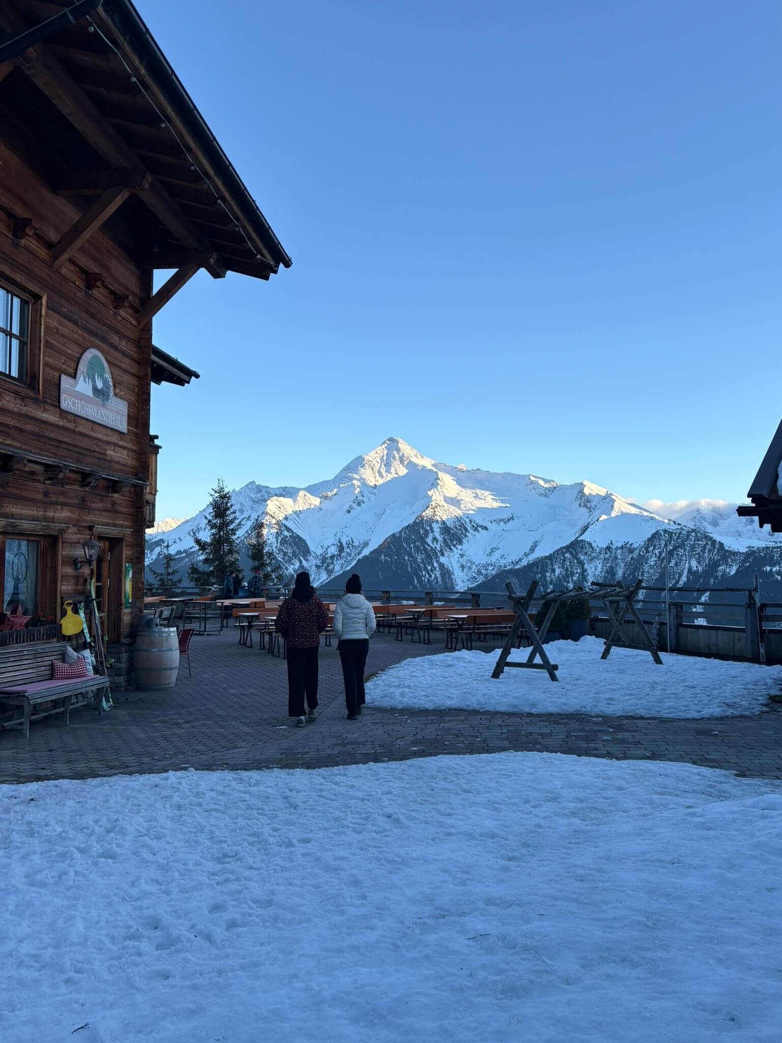 Zwei Personen gehen über die verschneite Terrasse einer Almhütte, gegenüber ein sonnenbeschienener Gipfel.