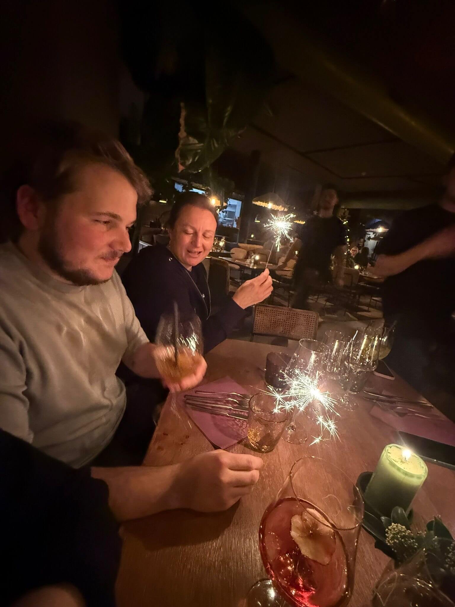 Teammitglieder stoßen mit Cocktails an, auf dem Tisch brennen Wunderkerzen und eine Kerze.