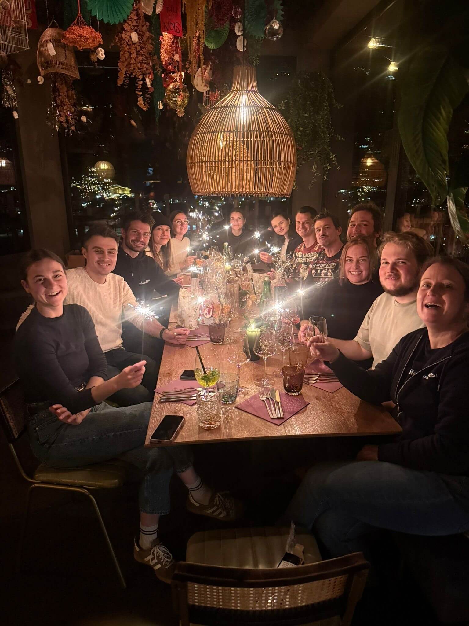 Das KLIXPERT-Team an einer langen Tafel im Restaurant, alle mit Wunderkerzen in der Hand, hinter den Fenstern die Lichter der Stadt bei Nacht.