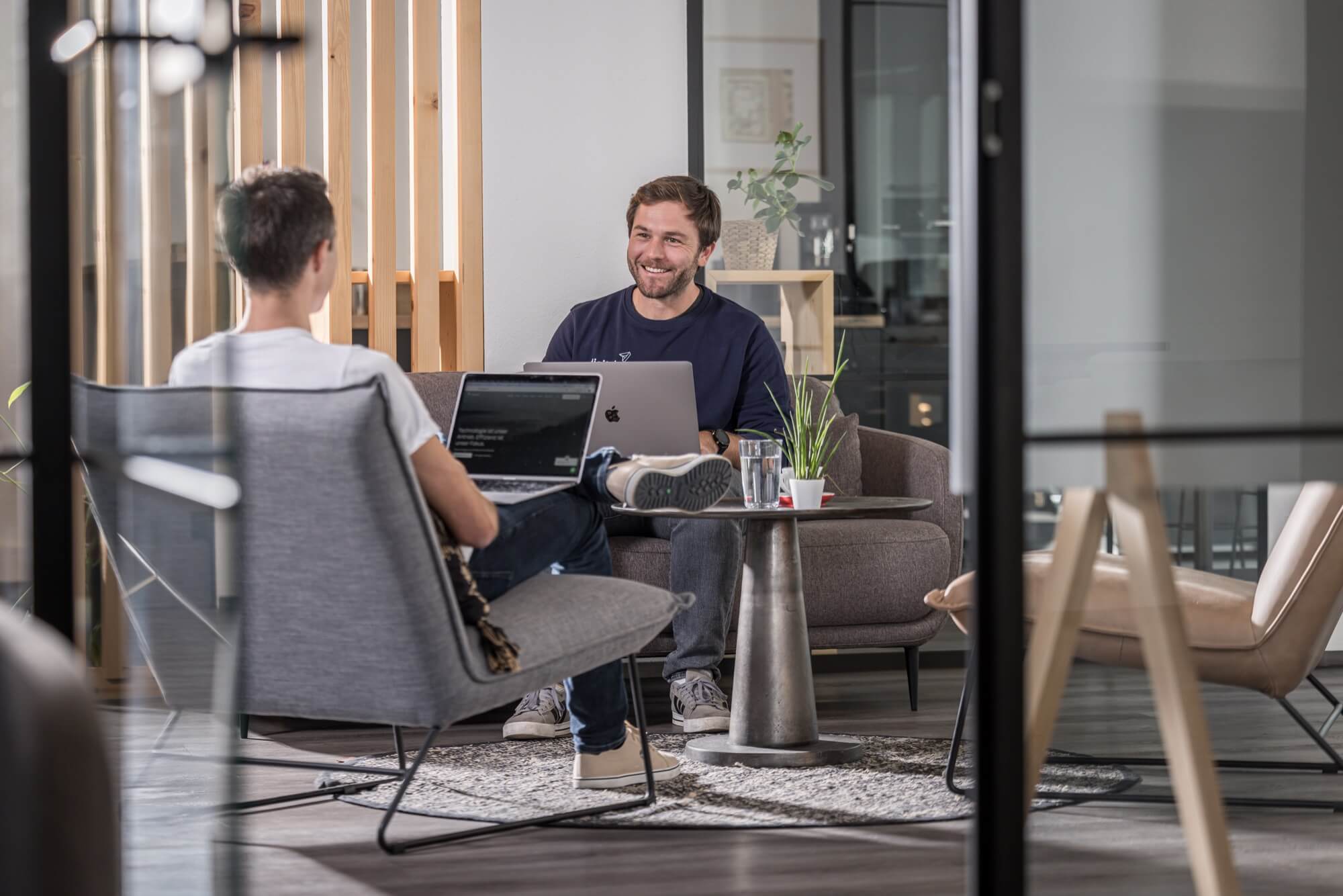 Zwei Männer in modernem Büro sitzen auf Sesseln, arbeiten an Laptops, lächeln und unterhalten sich.