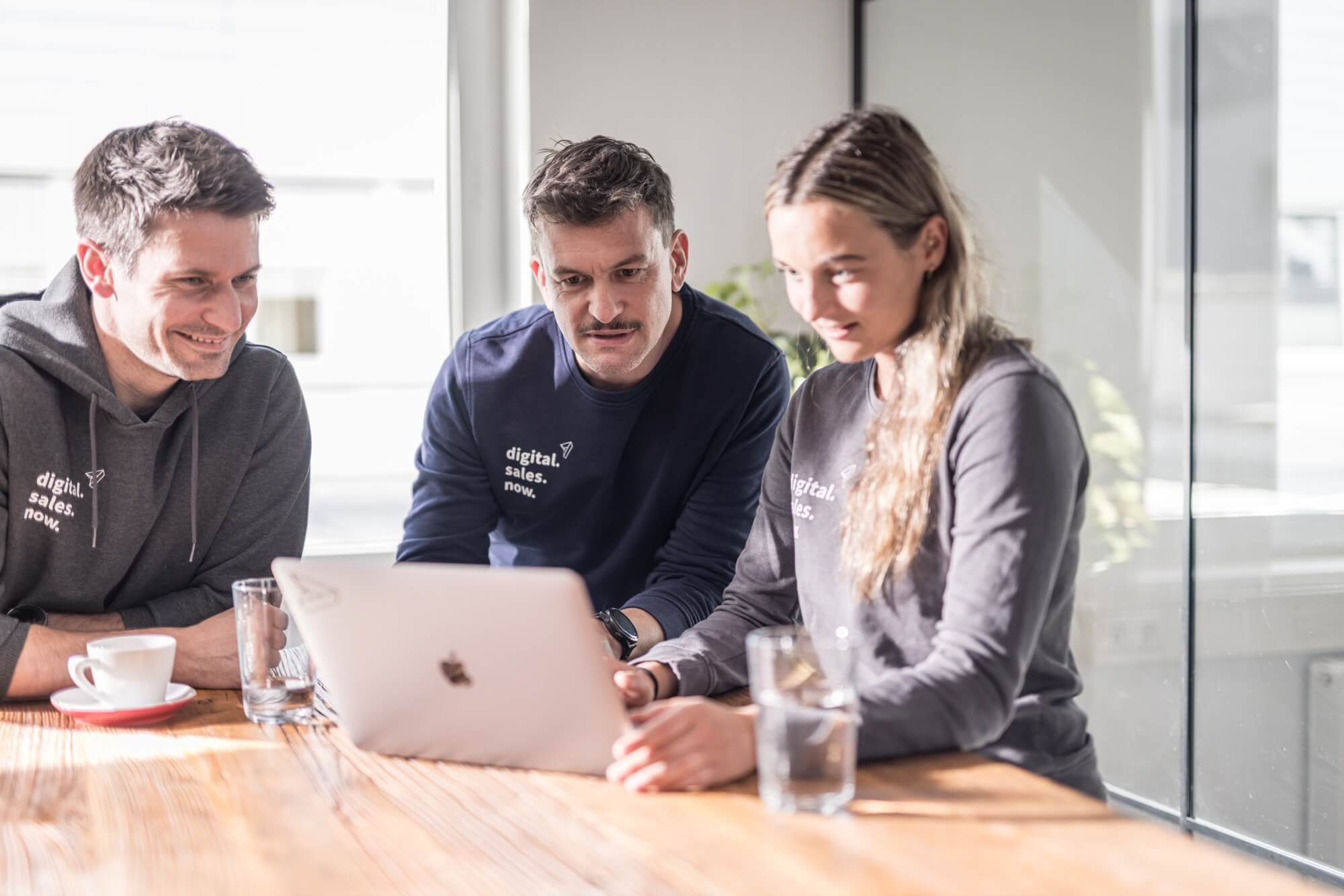 Drei Personen in einem Büro arbeiten konzentriert an einem Laptop. Sie tragen Pullover mit "digital. sales. now."