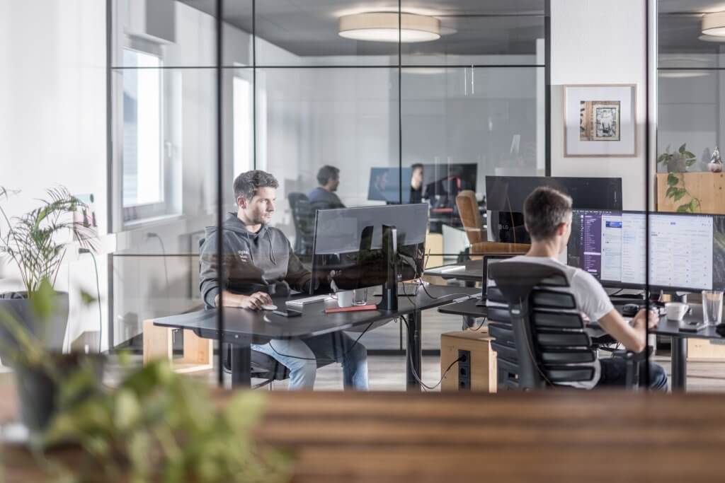 Männer arbeiten an Computern in modernem Büro mit Glaswänden.