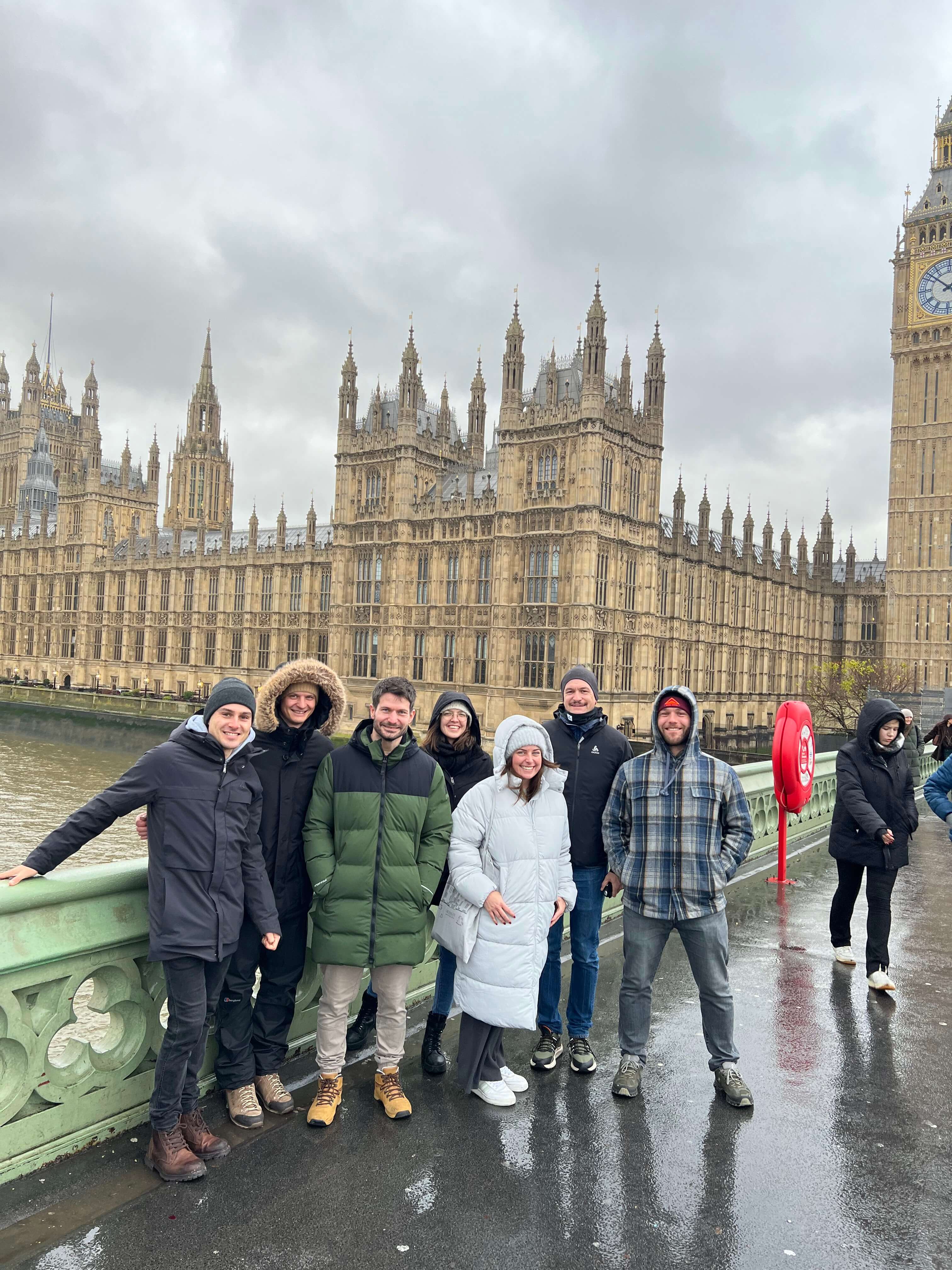 Gruppe von Menschen in Winterkleidung posiert vor dem Palace of Westminster in London, bewölkter Himmel.