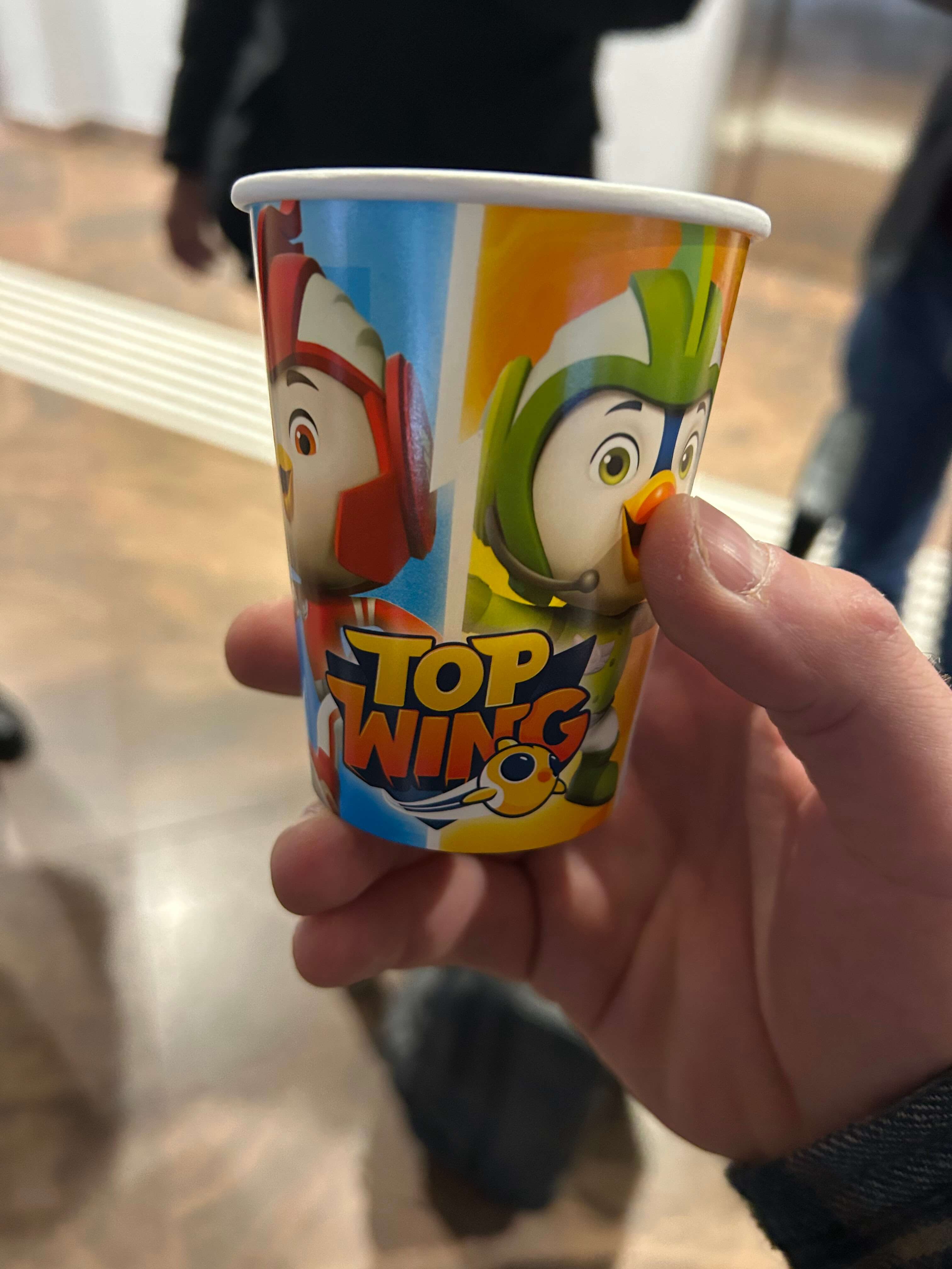 Hand hält einen bunten Pappbecher mit Top Wing-Cartoonfiguren in einer Innenumgebung.