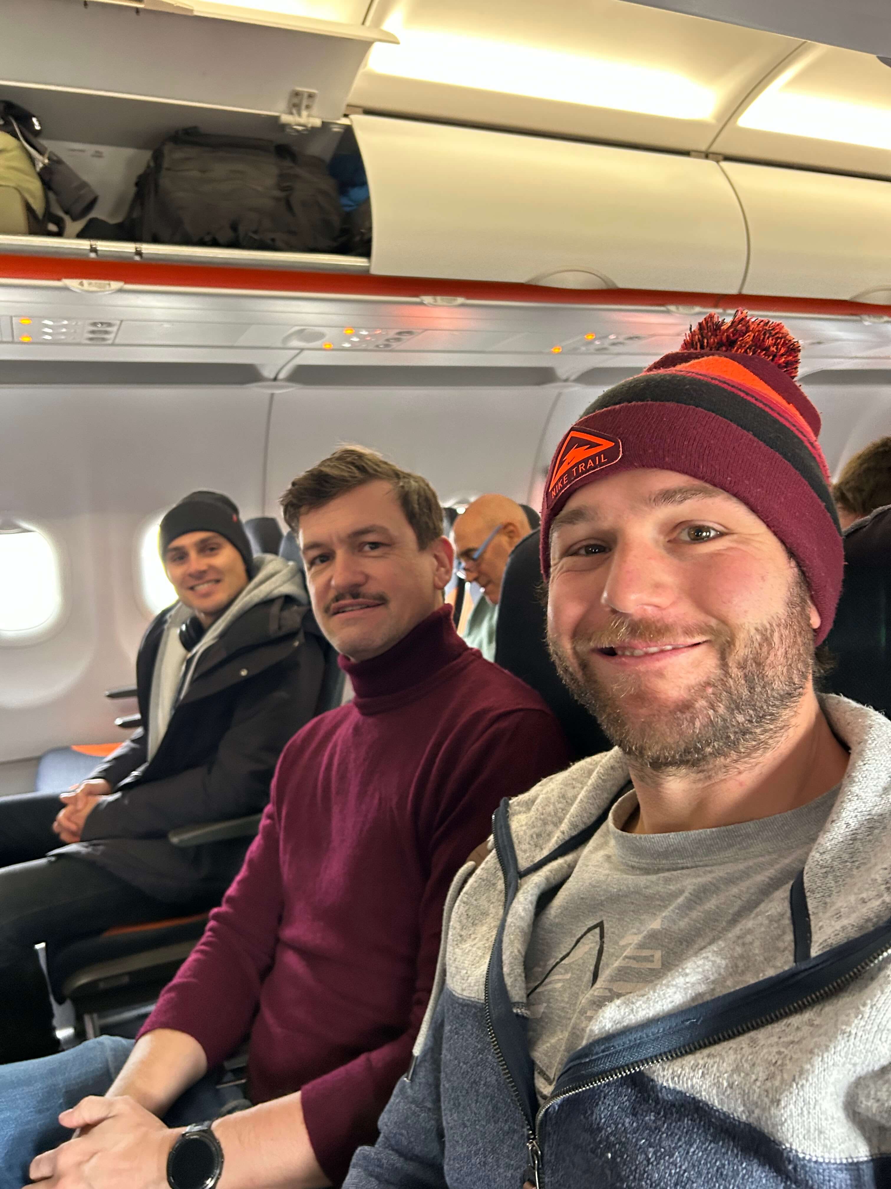 Drei Männer sitzen lächelnd in einer Flugzeugkabine, mit geöffneten Gepäckfächern über ihnen.