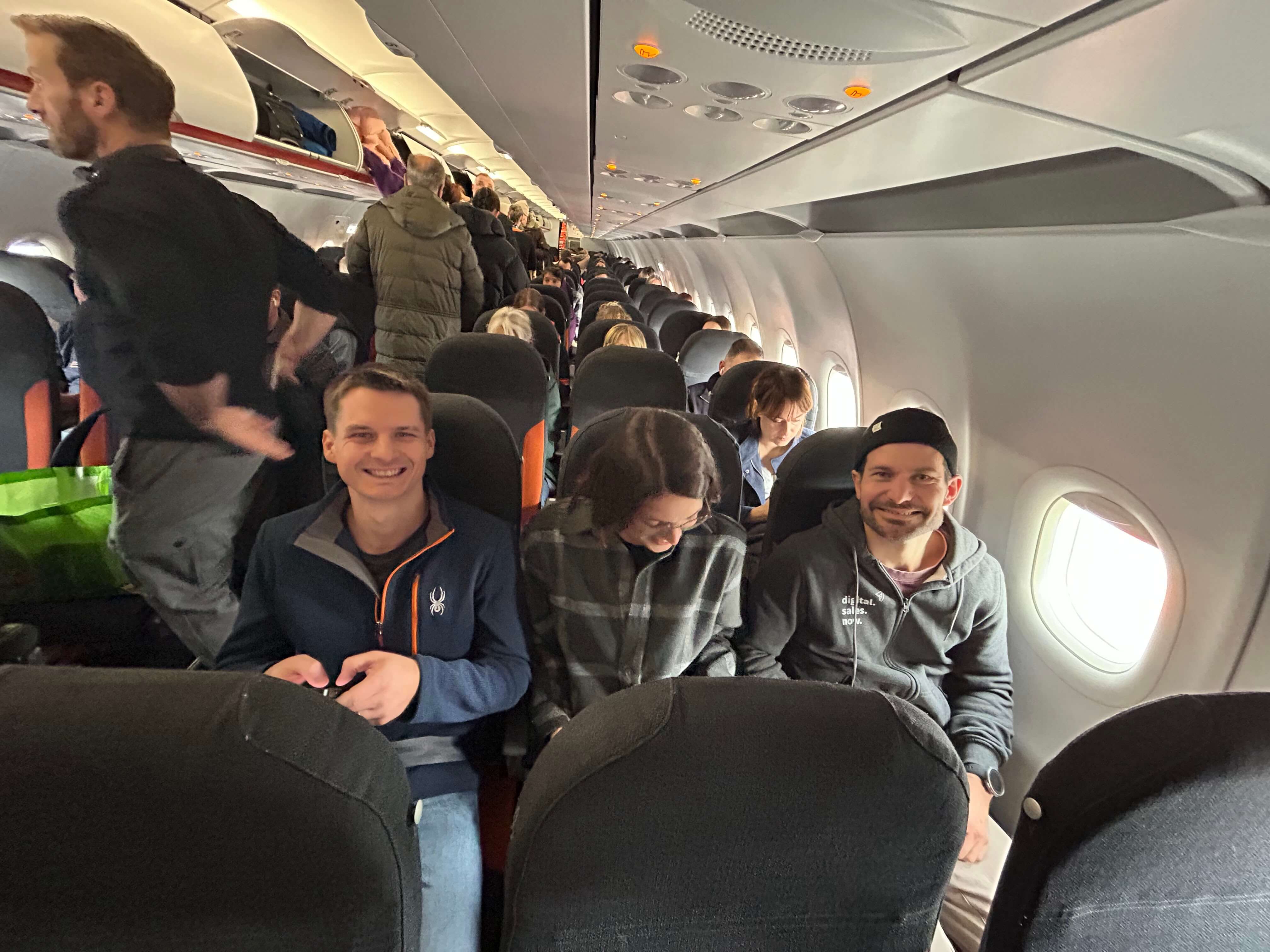 Passagiere sitzen in einem Flugzeug, während andere ihr Gepäck verstauen. Drei Personen lächeln in die Kamera.
