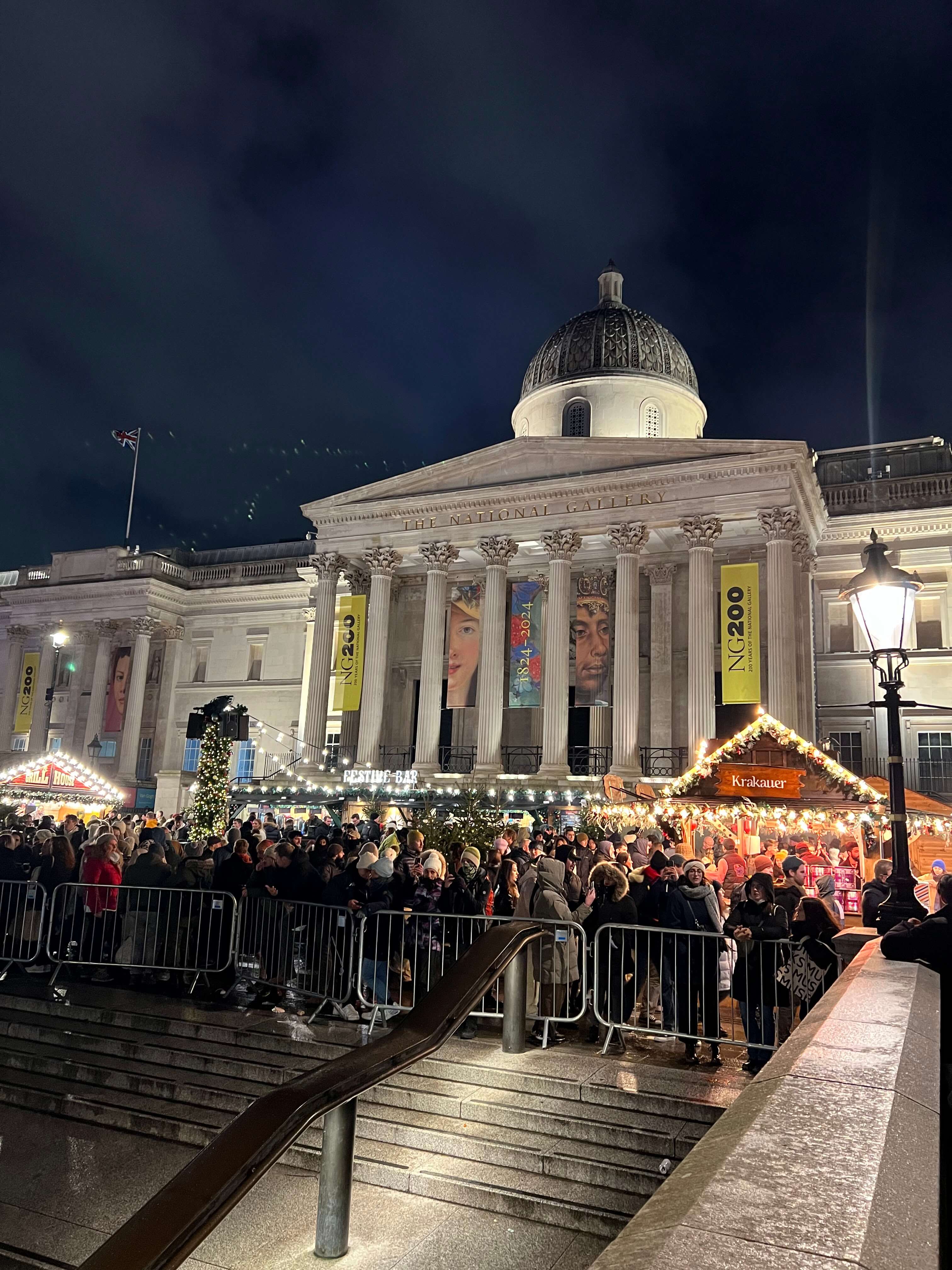 Menschenmenge vor beleuchteter National Gallery, London, bei Nacht. Weihnachtsmarktstände und Dekorationen im Vordergrund.