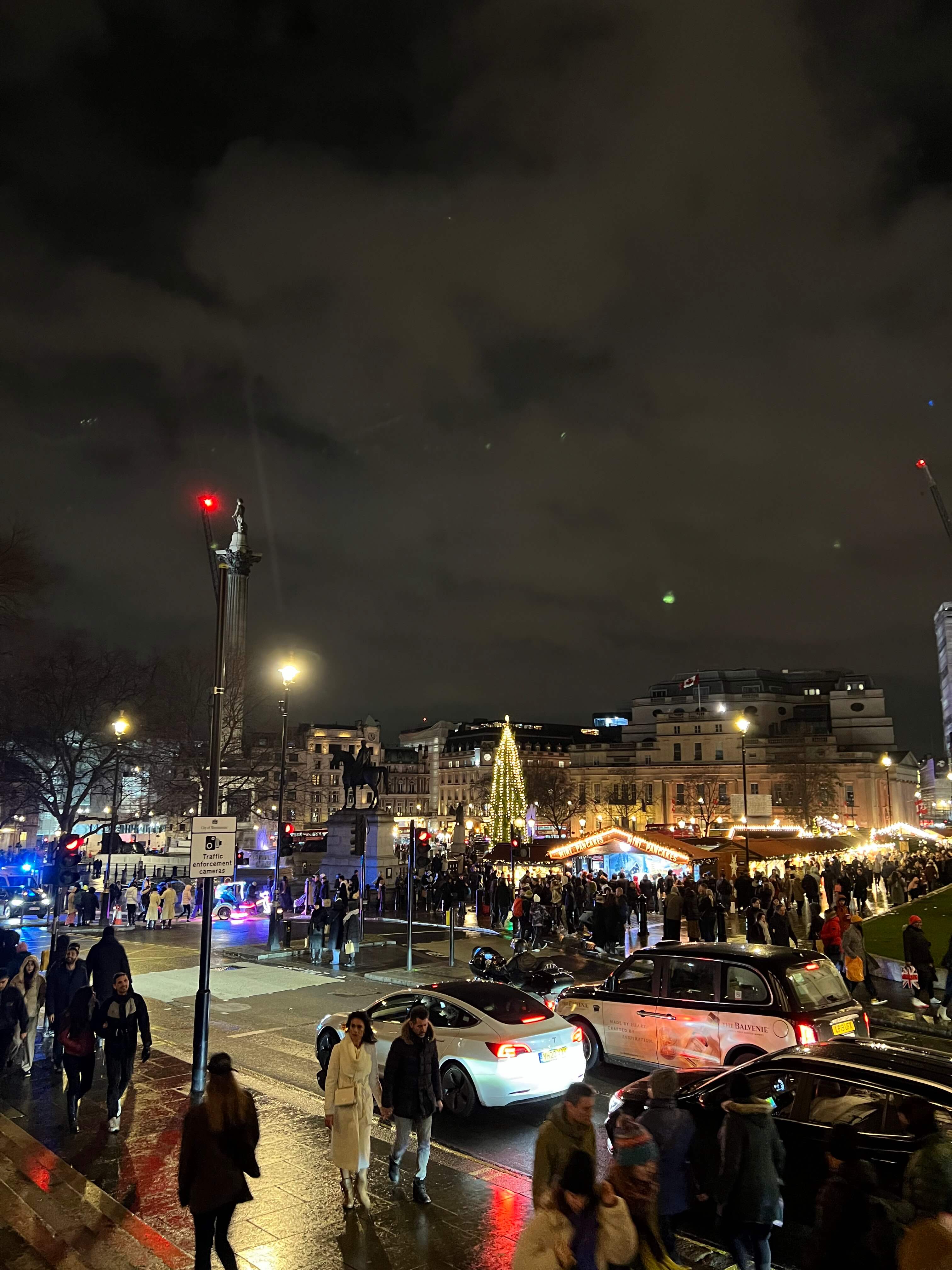 Nachtaufnahme eines belebten Weihnachtsmarktes mit beleuchtetem Baum, vielen Besuchern und Autos auf der Straße.