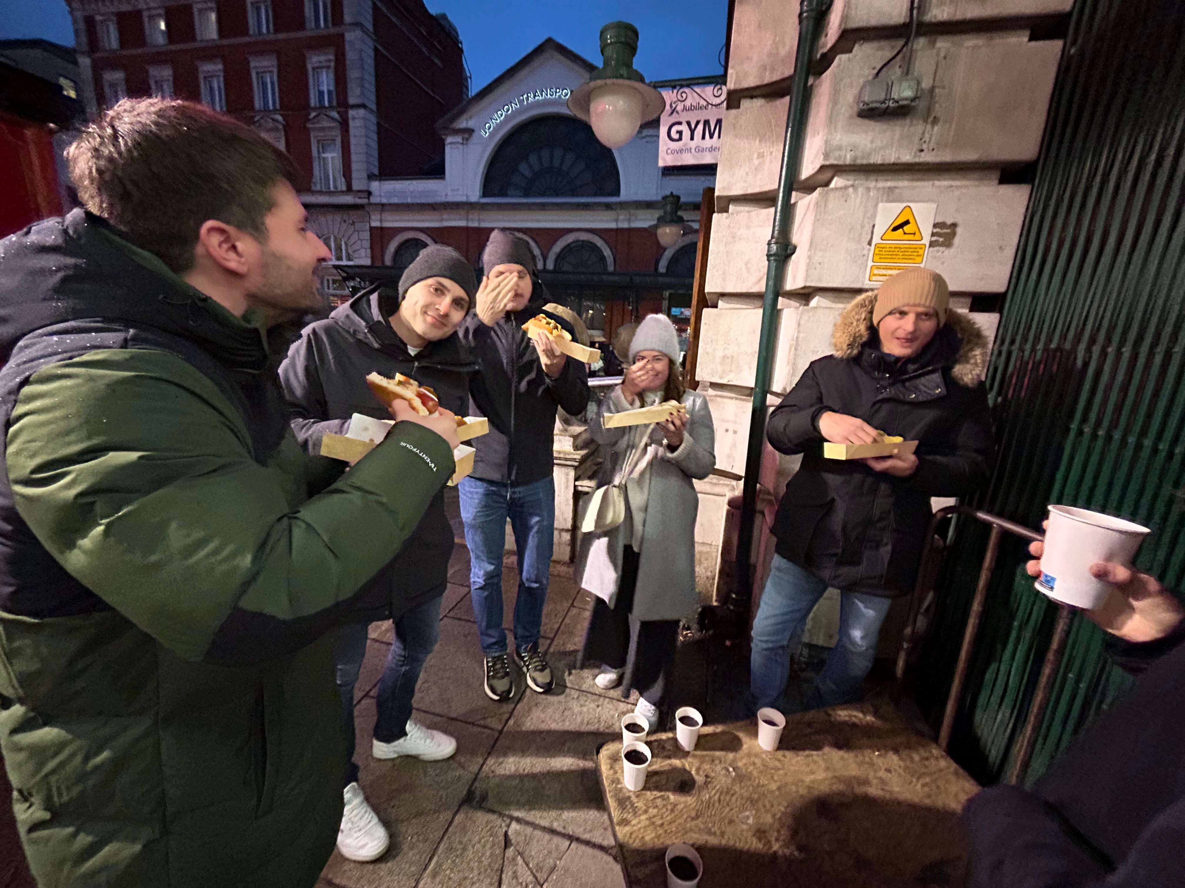 Gruppe von Menschen isst Hotdogs vor einem Gebäude in der Stadt bei Abenddämmerung.
