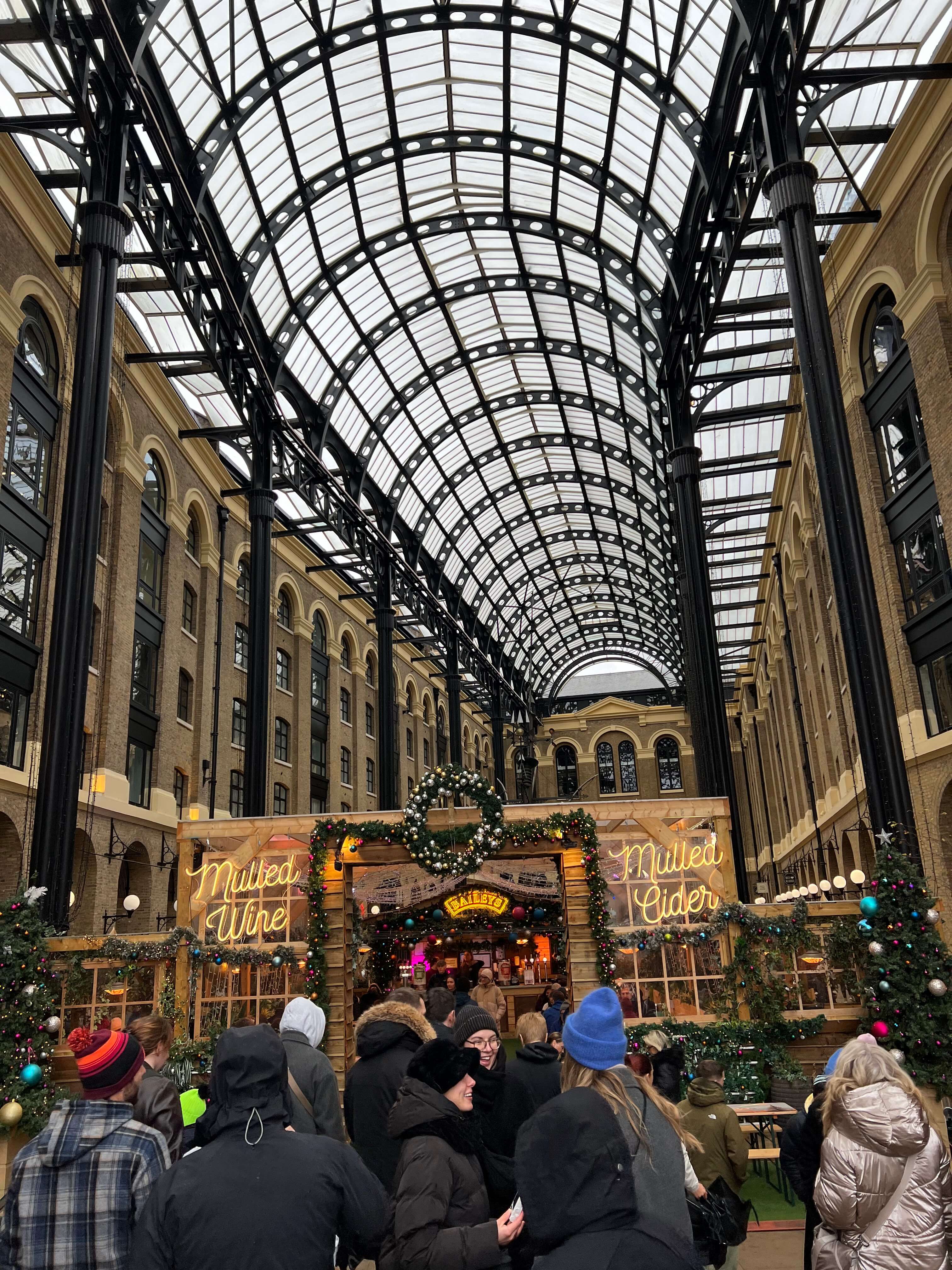 Weihnachtsmarkt in überdachter Passage mit Lichterketten, Menschen in Winterkleidung, Schilder für Glühwein und Cider.