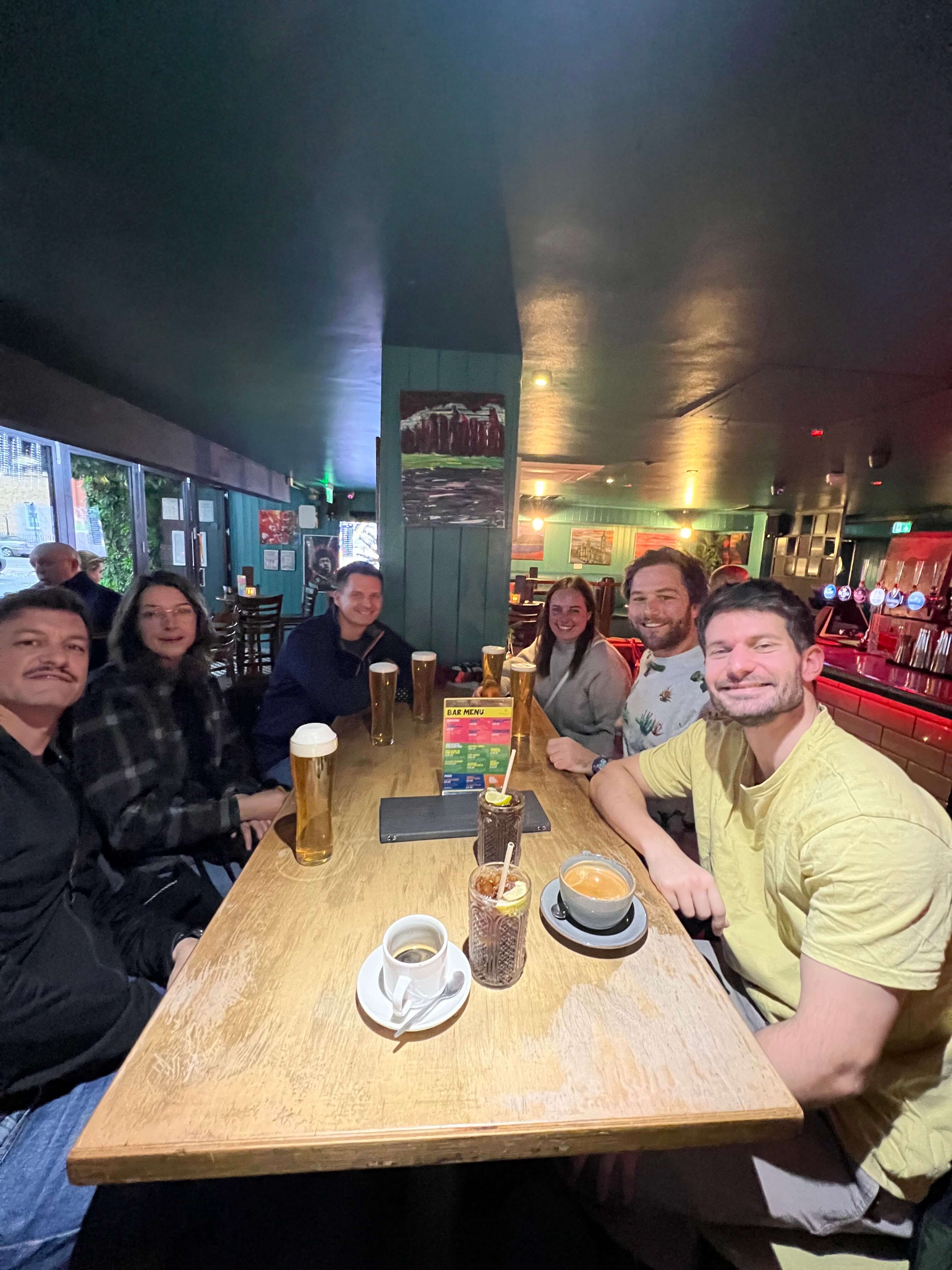 Gruppe von sechs Personen sitzt in einer Bar an einem Tisch mit Getränken und lächelt in die Kamera.