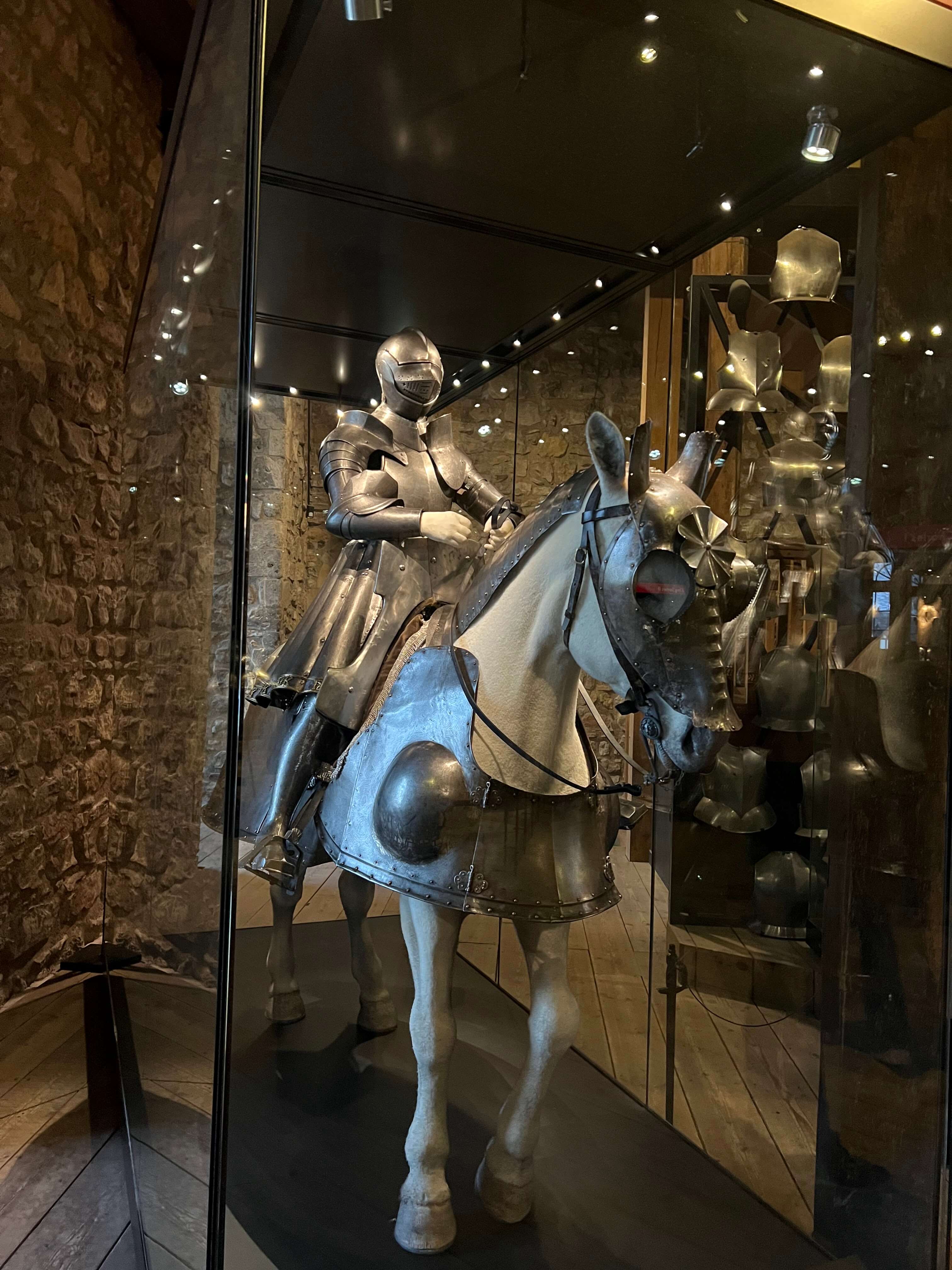 Ritterrüstung auf einem Pferd in einem Museum ausgestellt, umgeben von Rüstungen und Steinwänden.