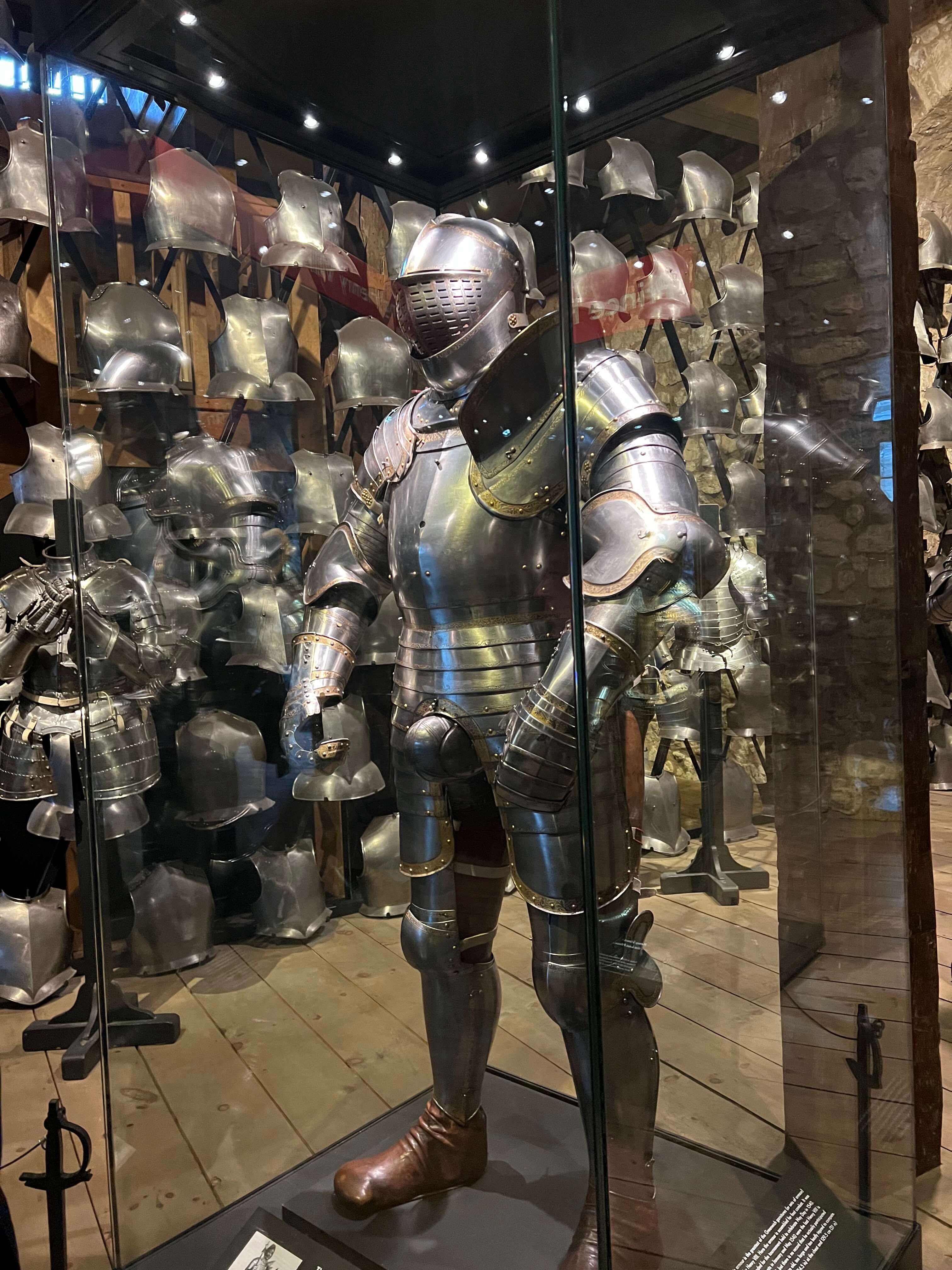 Ritterrüstung in Glasvitrine, umgeben von weiteren Helmen und Rüstungen in einem Museum.