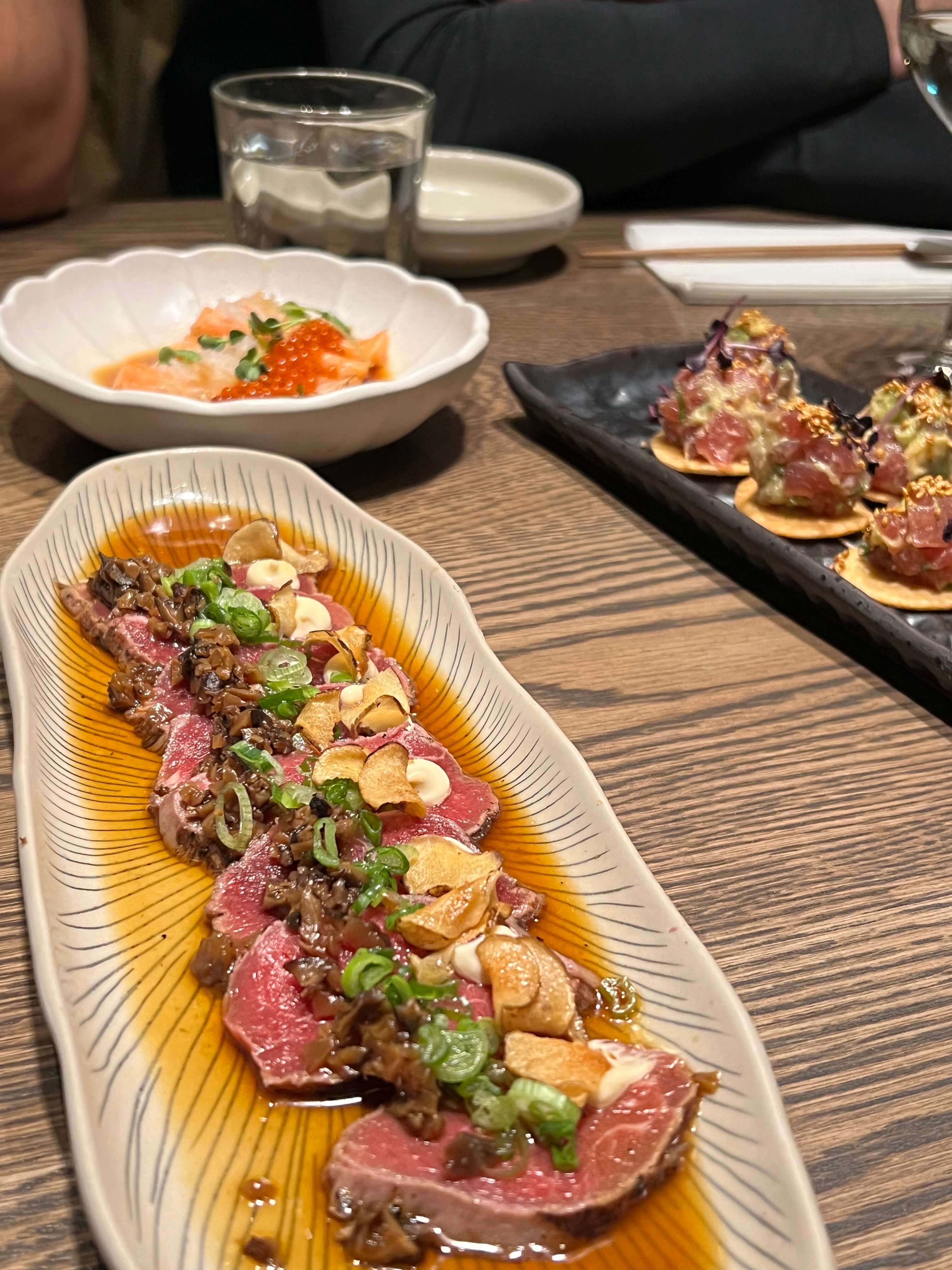 Japanisches Gericht mit rohem Fleisch, Frühlingszwiebeln und Knoblauchchips auf Teller, im Hintergrund Lachs und Avocado-Tatar.