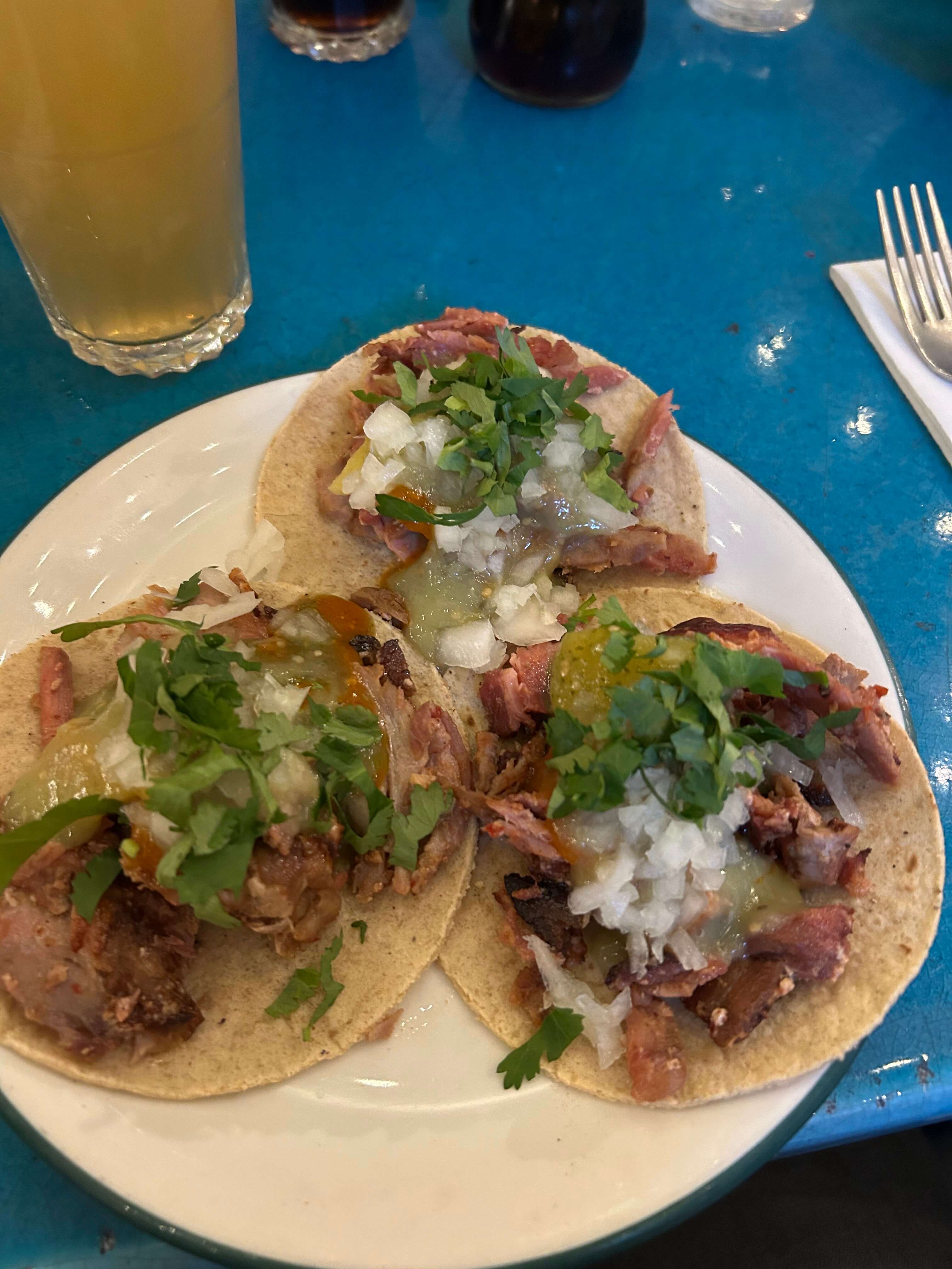 Drei Tacos mit Fleisch, Zwiebeln, Koriander und Salsa auf einem Teller, daneben ein Glas Getränk.
