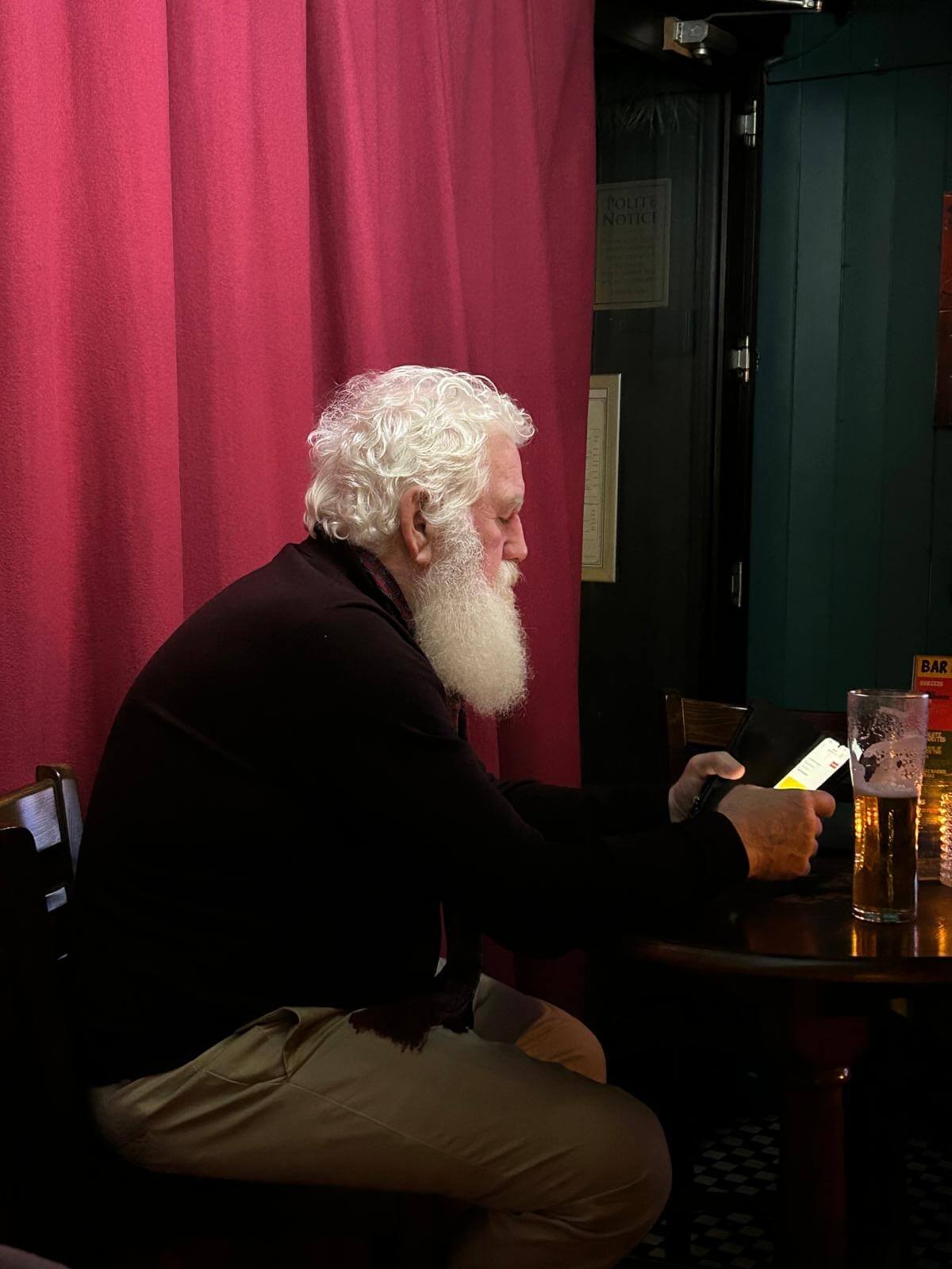 Älterer Mann mit weißem Bart sitzt in einer Bar und liest auf seinem Smartphone. Neben ihm ein volles Bierglas.