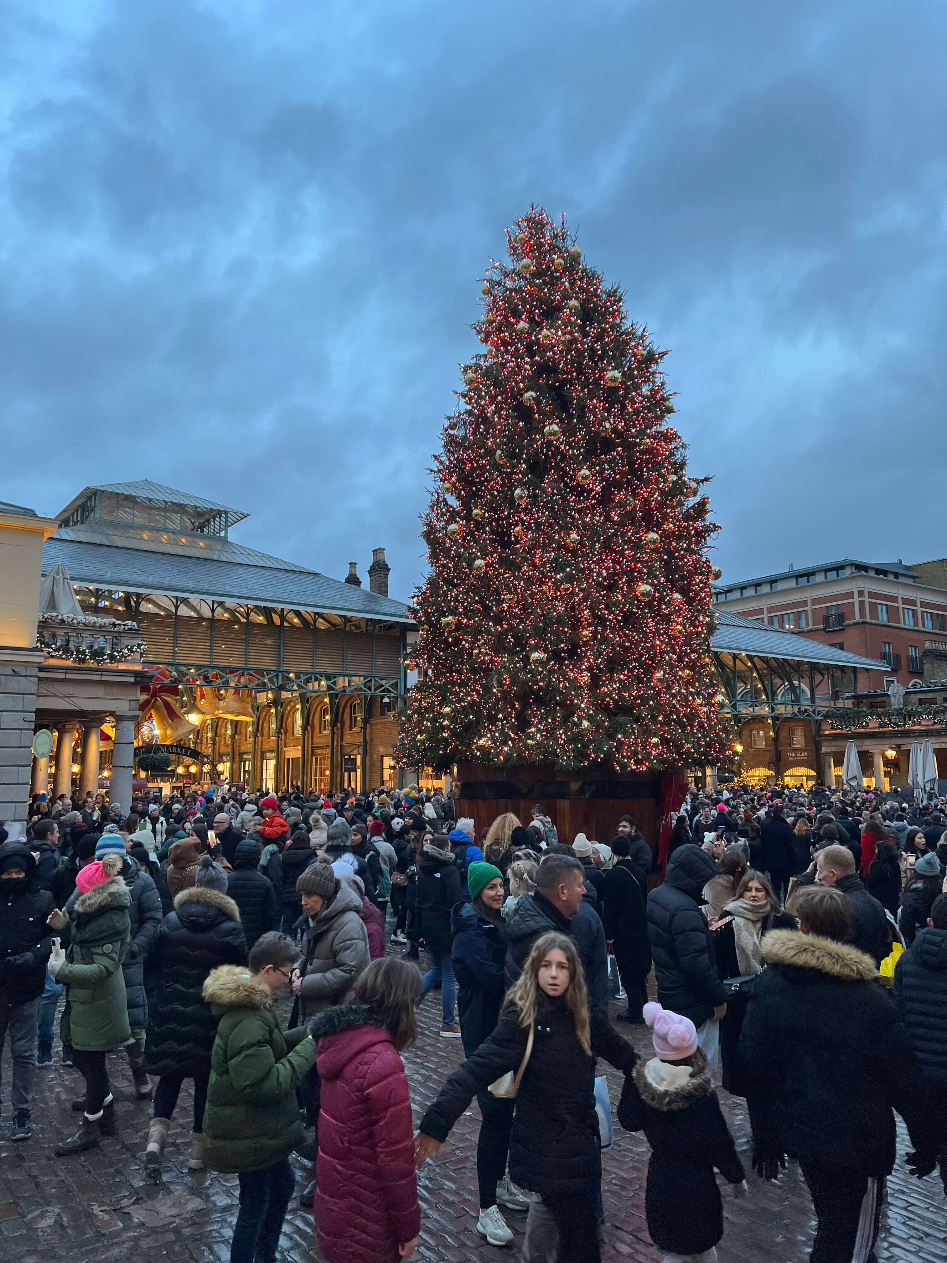 Großer, beleuchteter Weihnachtsbaum auf einem belebten Platz mit vielen Menschen bei Abenddämmerung.